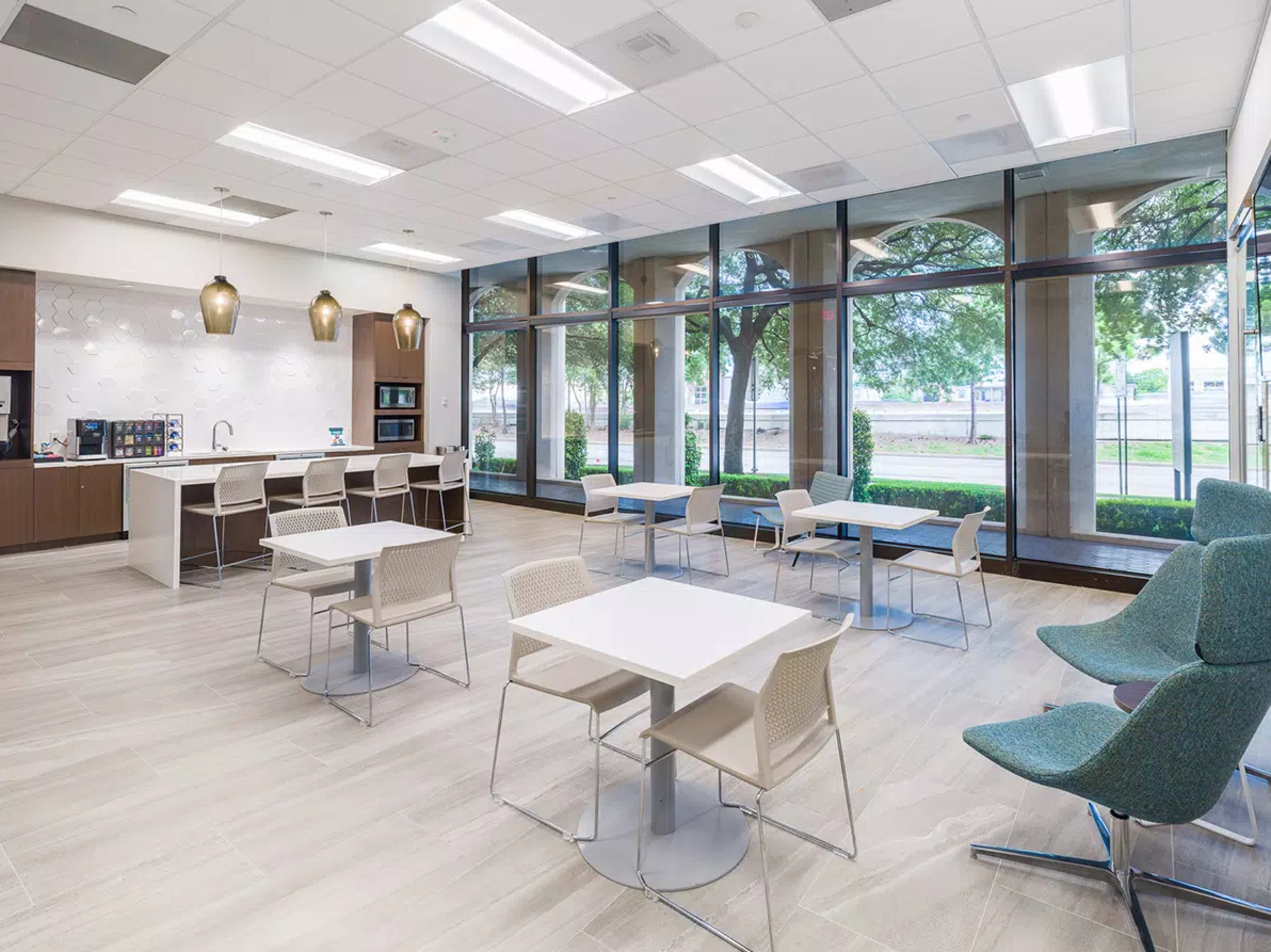 The image shows a modern, bright communal area with a kitchenette, seating arrangements, and large windows that overlook an outdoor space.