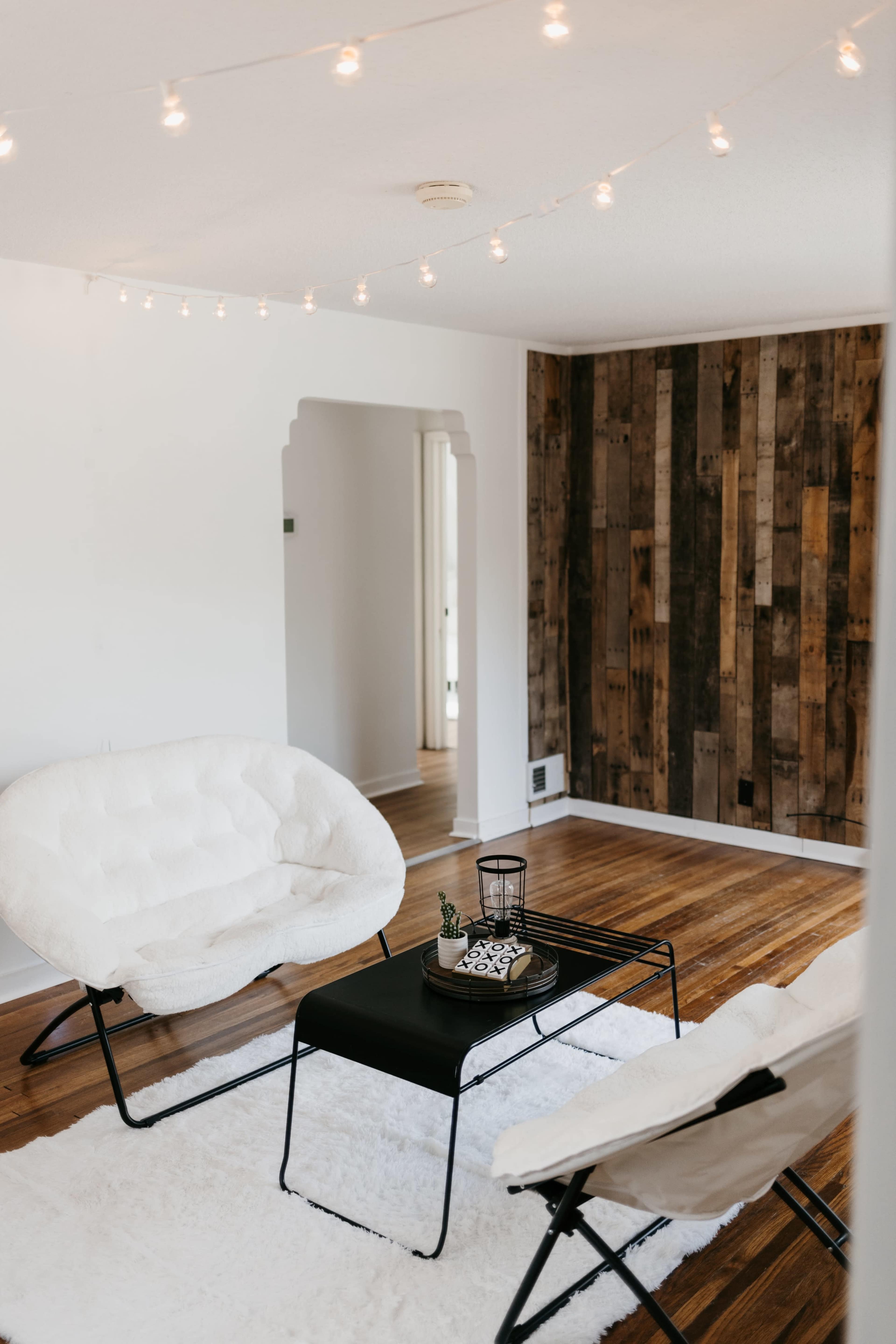 A living room with two white chairs, a black table, and wood-paneled walls, illuminated by string lights overhead.