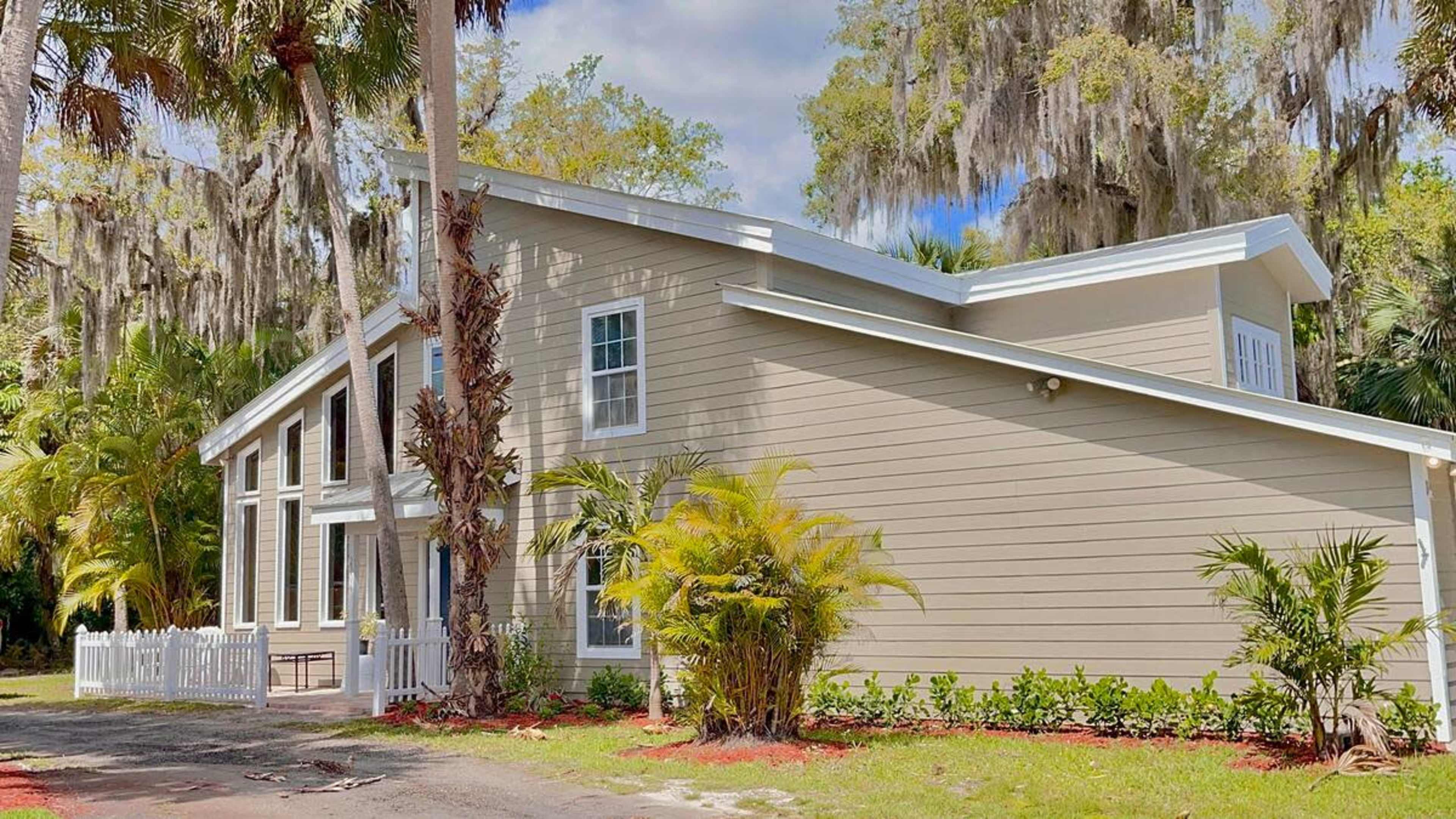 A light-colored house with large windows and palm trees, located in a landscaped area with a gravel path.