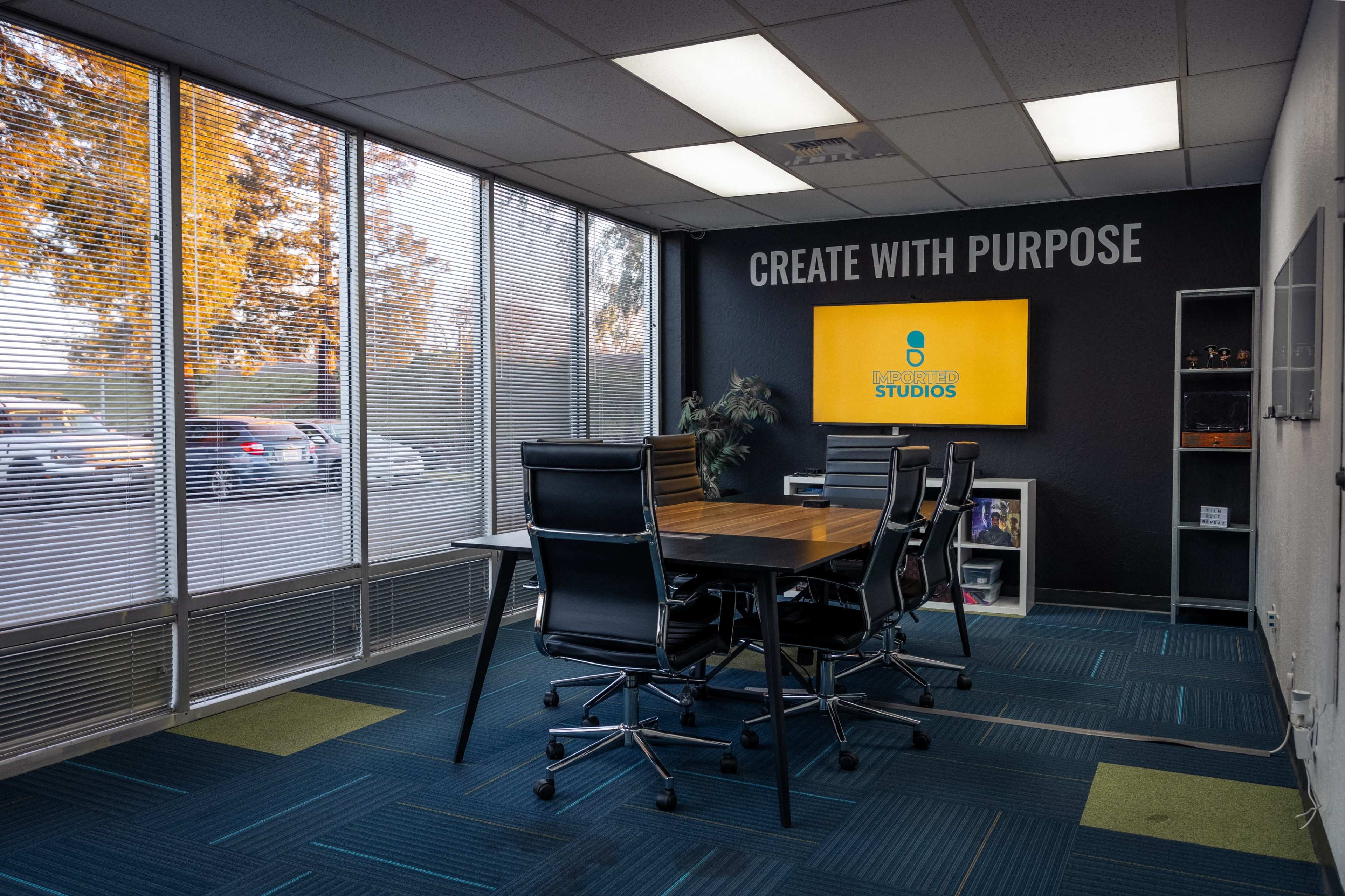 A conference room features a table surrounded by black chairs, large windows letting in daylight, and a wall with the phrase "CREATE WITH PURPOSE" and a screen displaying the name "STUDIOS."