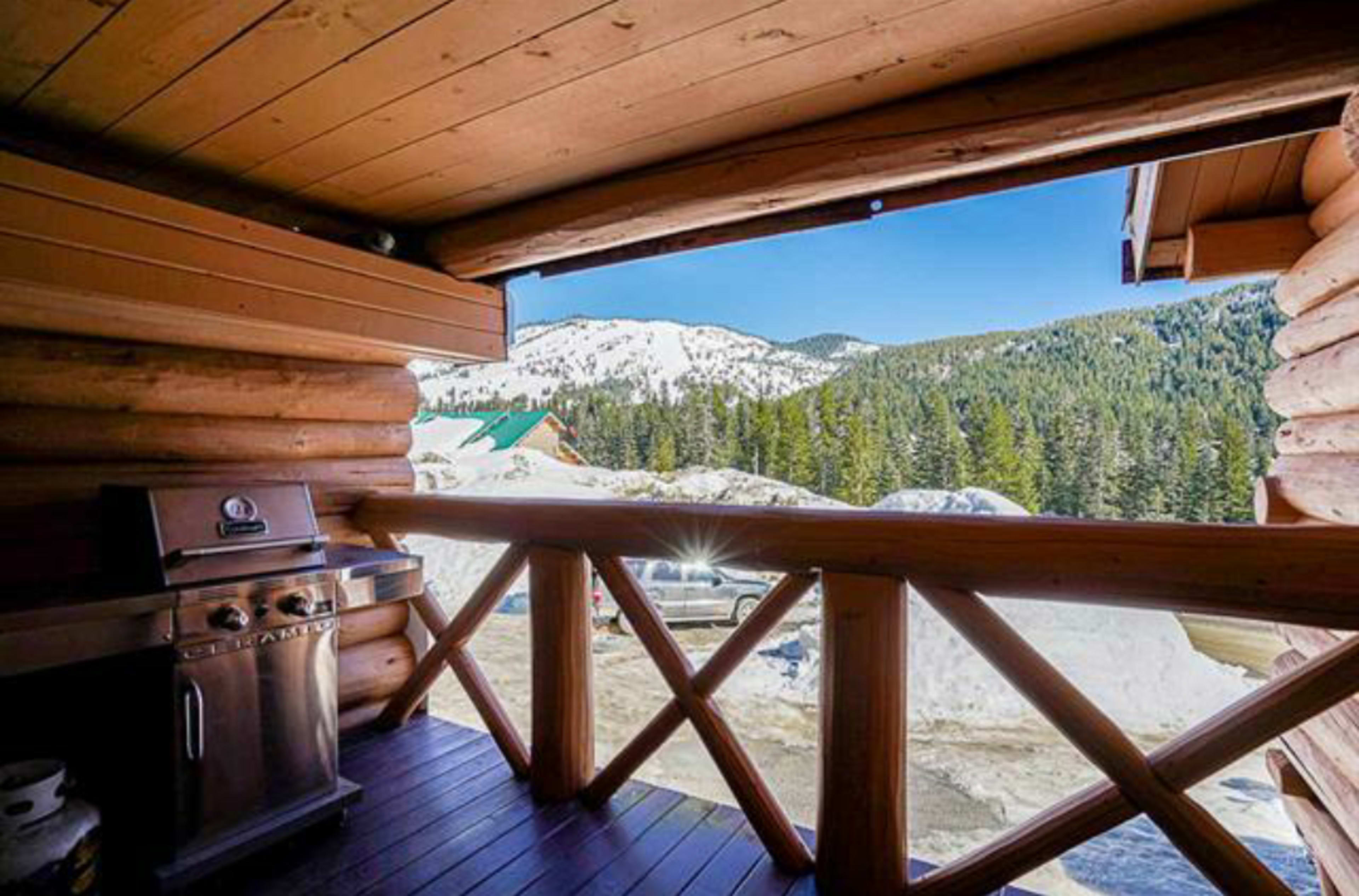 A wooden balcony features a grill overlooking a mountainous landscape with snow-covered peaks and dense forests.