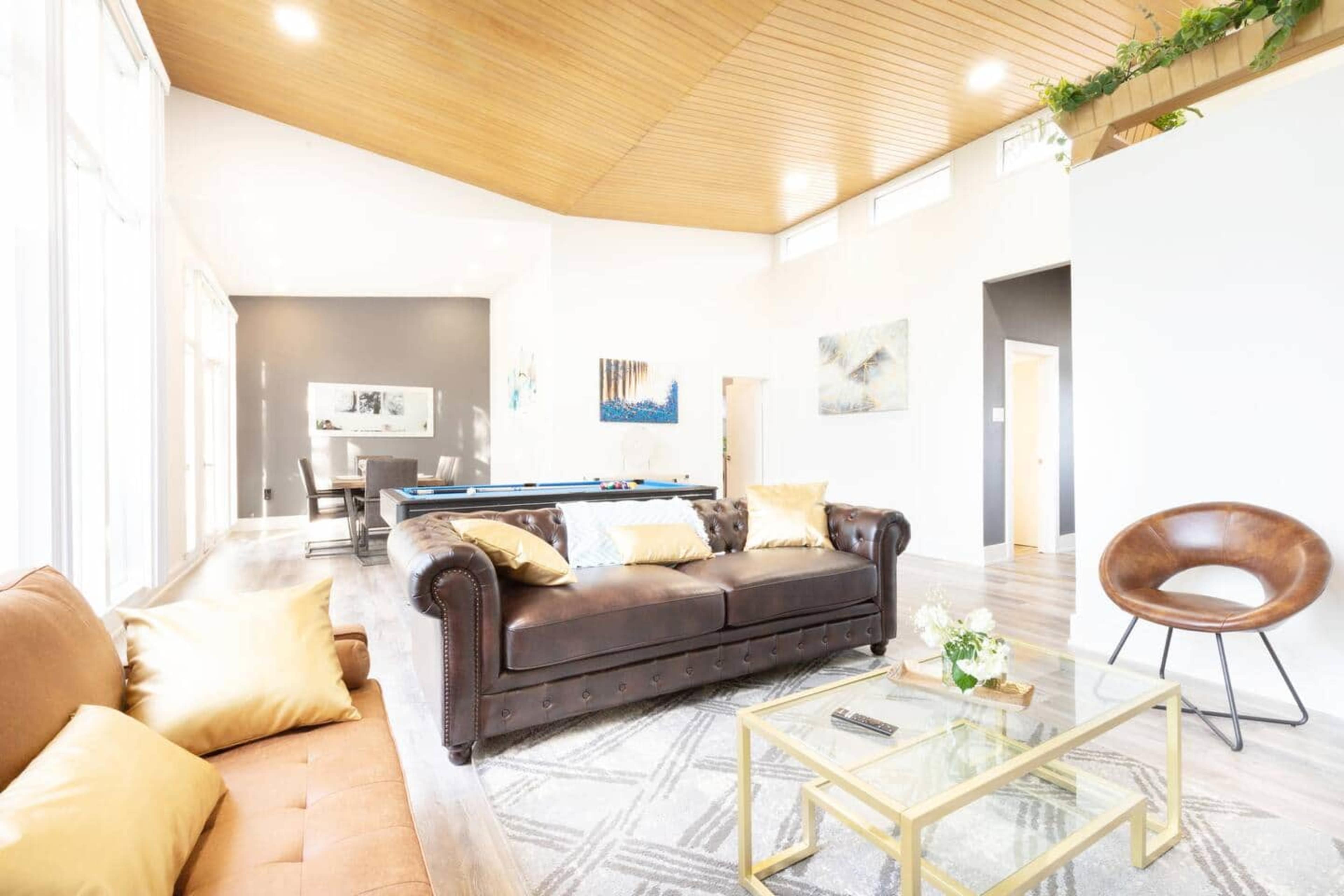 A spacious living area features a brown leather sofa, a glass coffee table, and large windows that provide natural light.