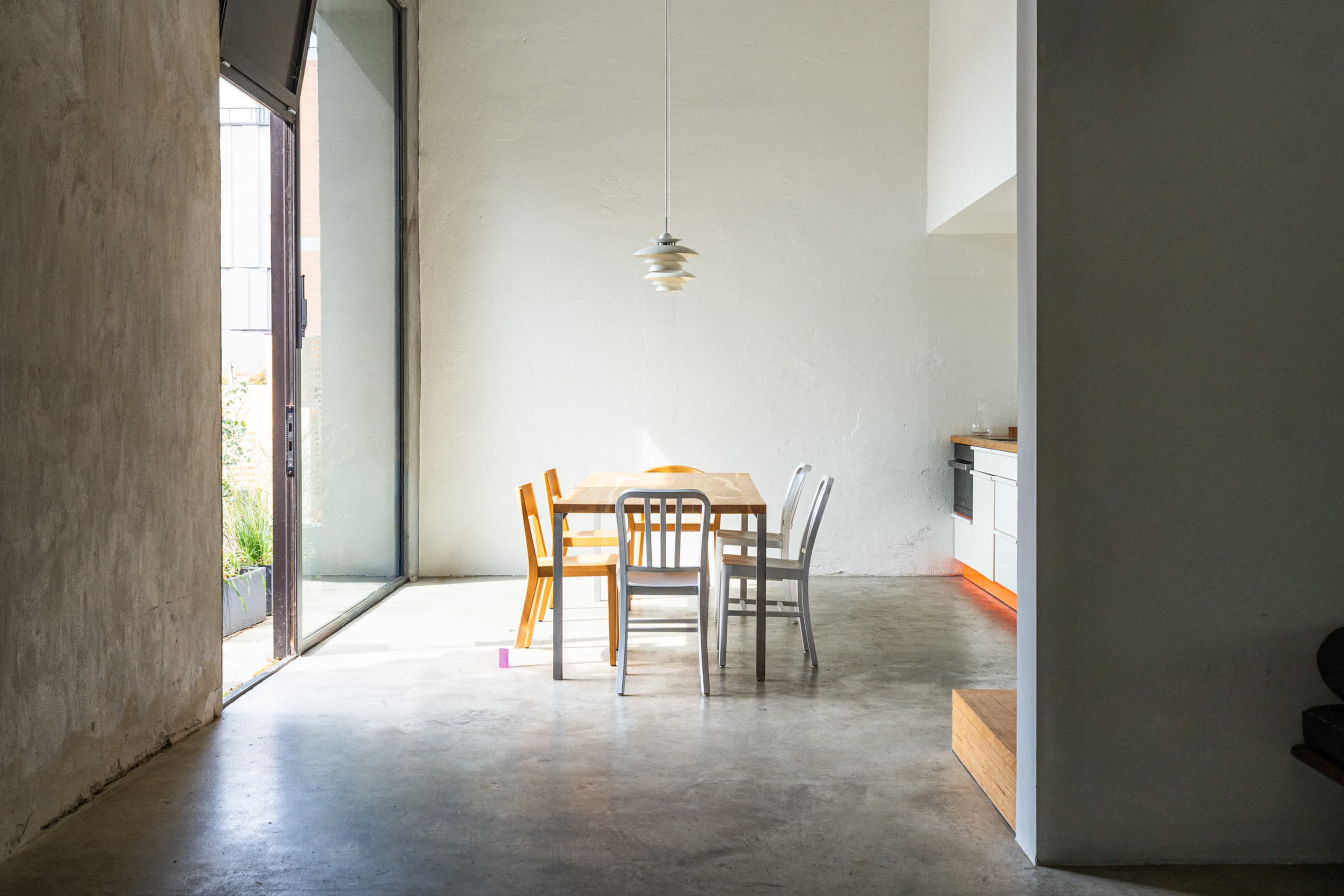 A minimalist dining area features a wooden table surrounded by four chairs of varying colors, illuminated by natural light from a large window.