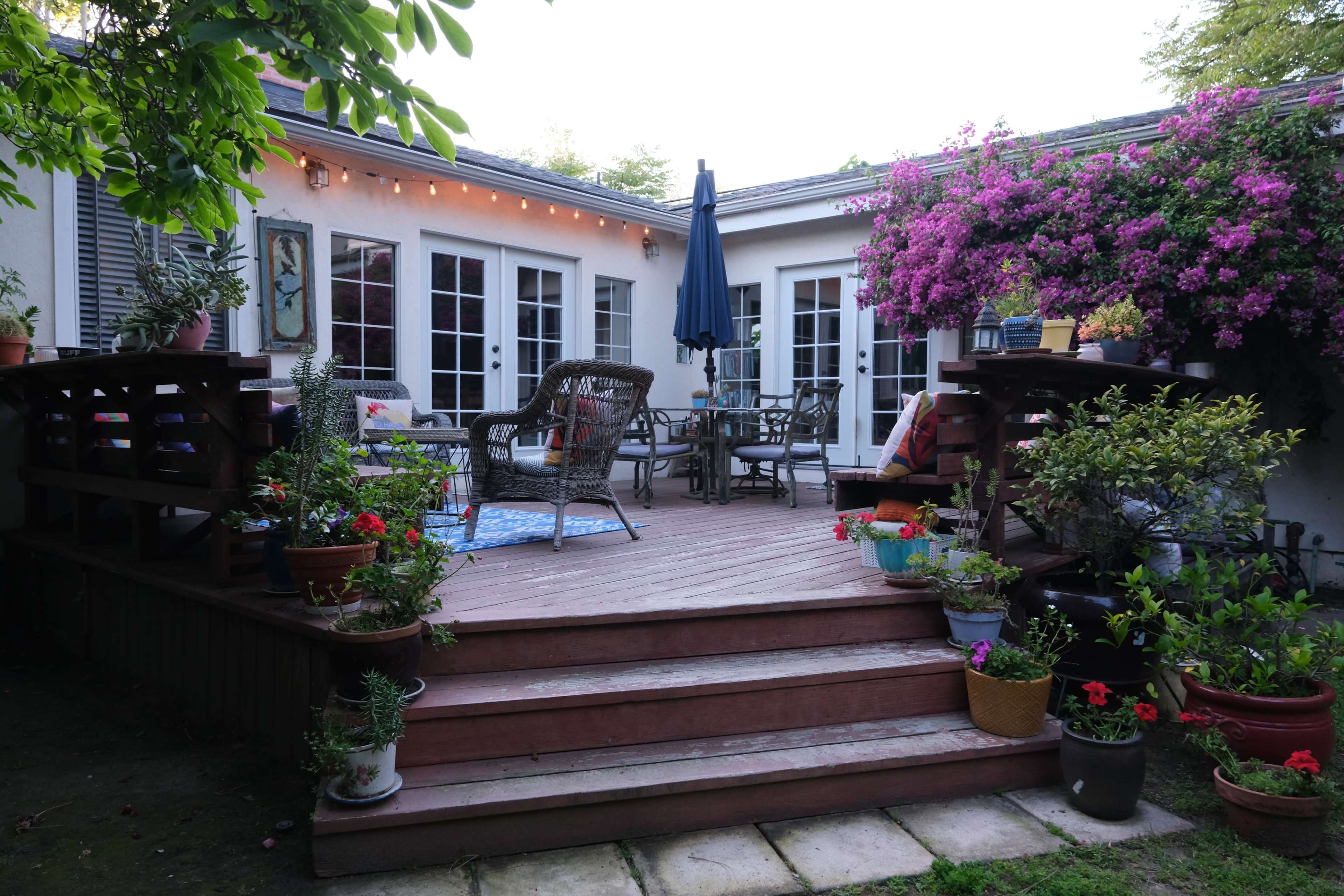 A wooden deck surrounded by potted flowers and plants, leading to a seating area with a table and chairs, illuminated by string lights.