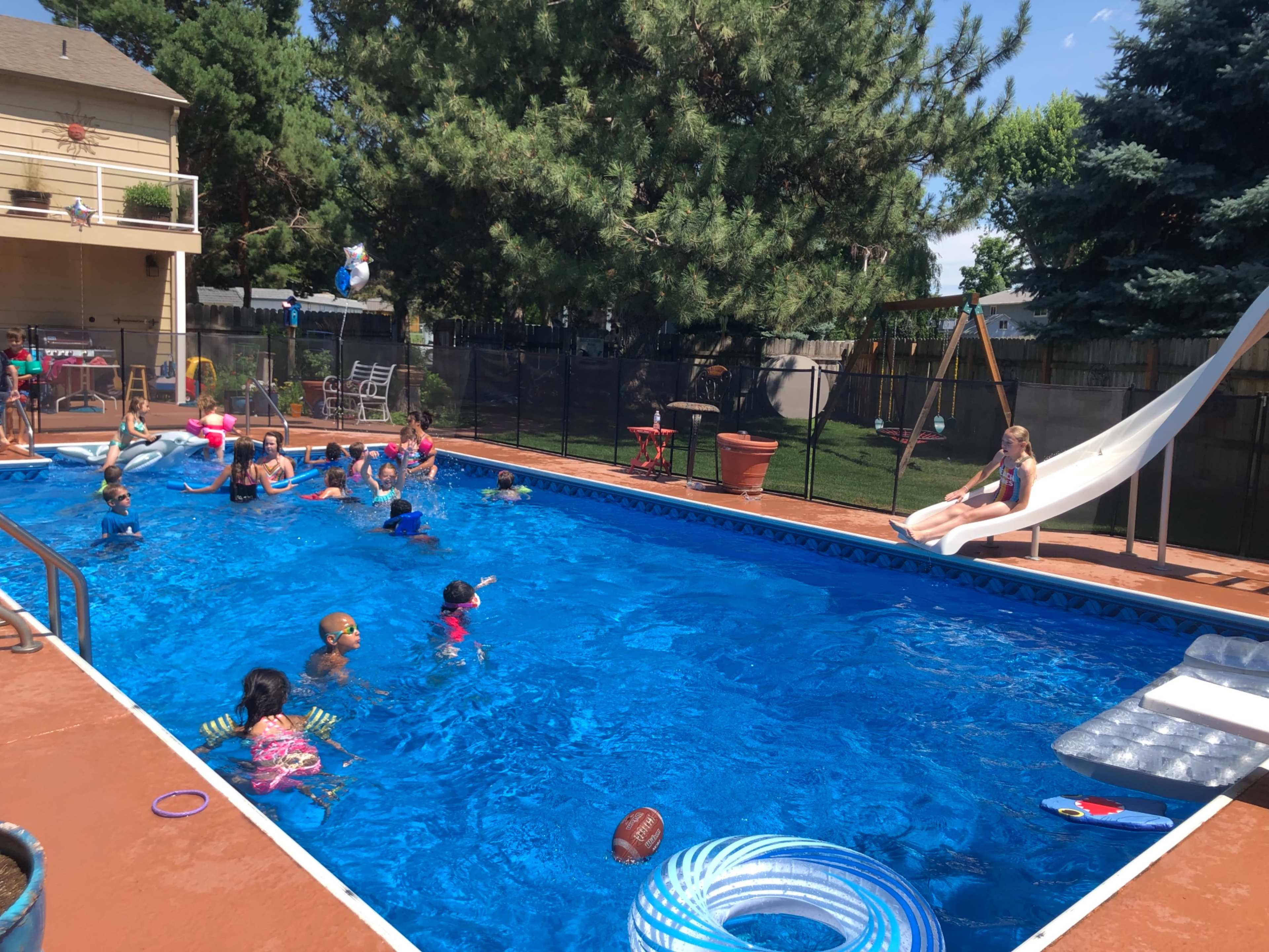 A group of children swims and plays in a blue pool while one child sits on a slide overlooking the water.