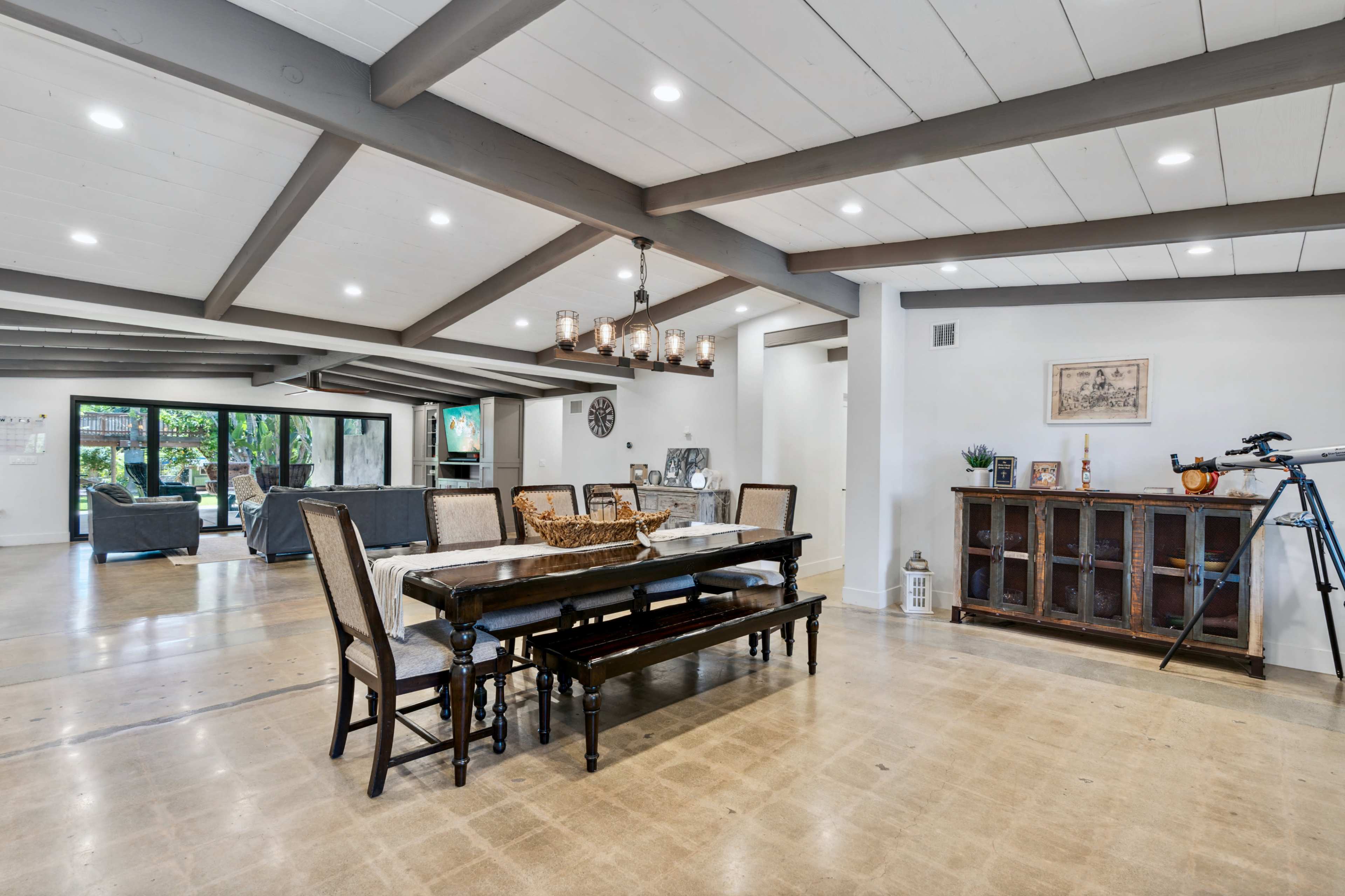 An open-concept dining area featuring a long wooden table with chairs, surrounded by a living space with a seating area, all illuminated by natural light.