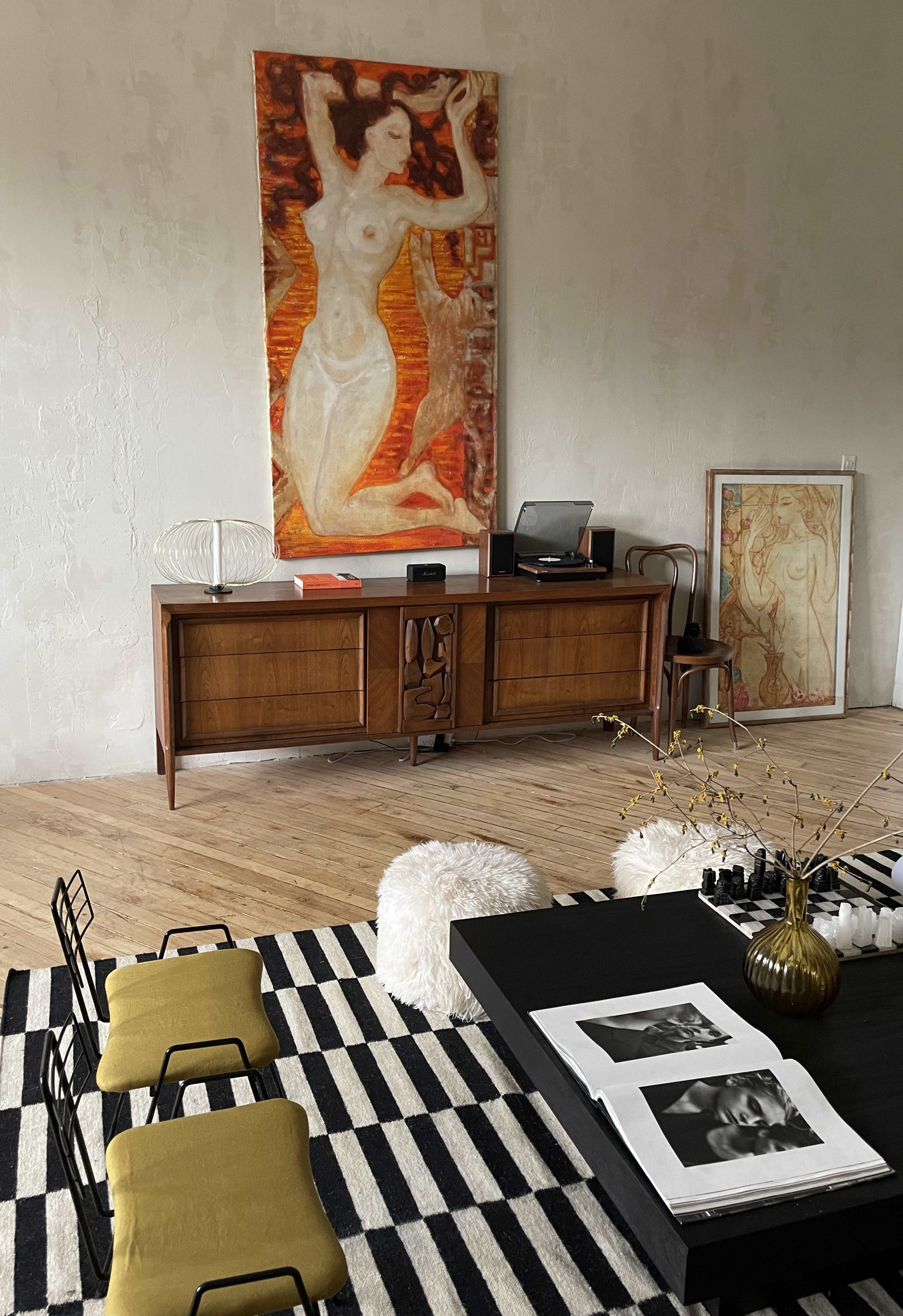 The image features a stylish interior with a wooden sideboard, an abstract painting of a nude woman on the wall, and a coffee table with magazines, set against a striped rug.