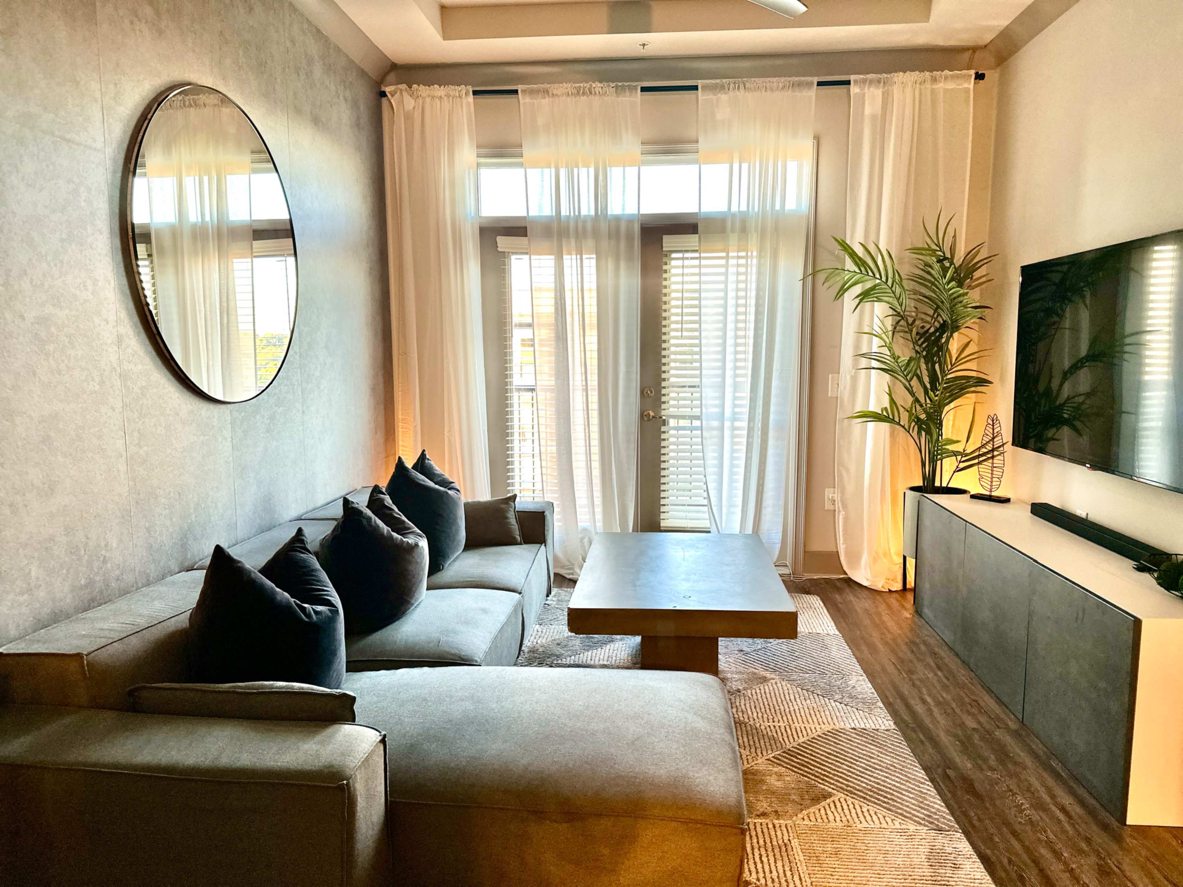 A modern living room features a sectional sofa, a round mirror, large windows with sheer curtains, a flat-screen TV on a cabinet, and a potted plant.