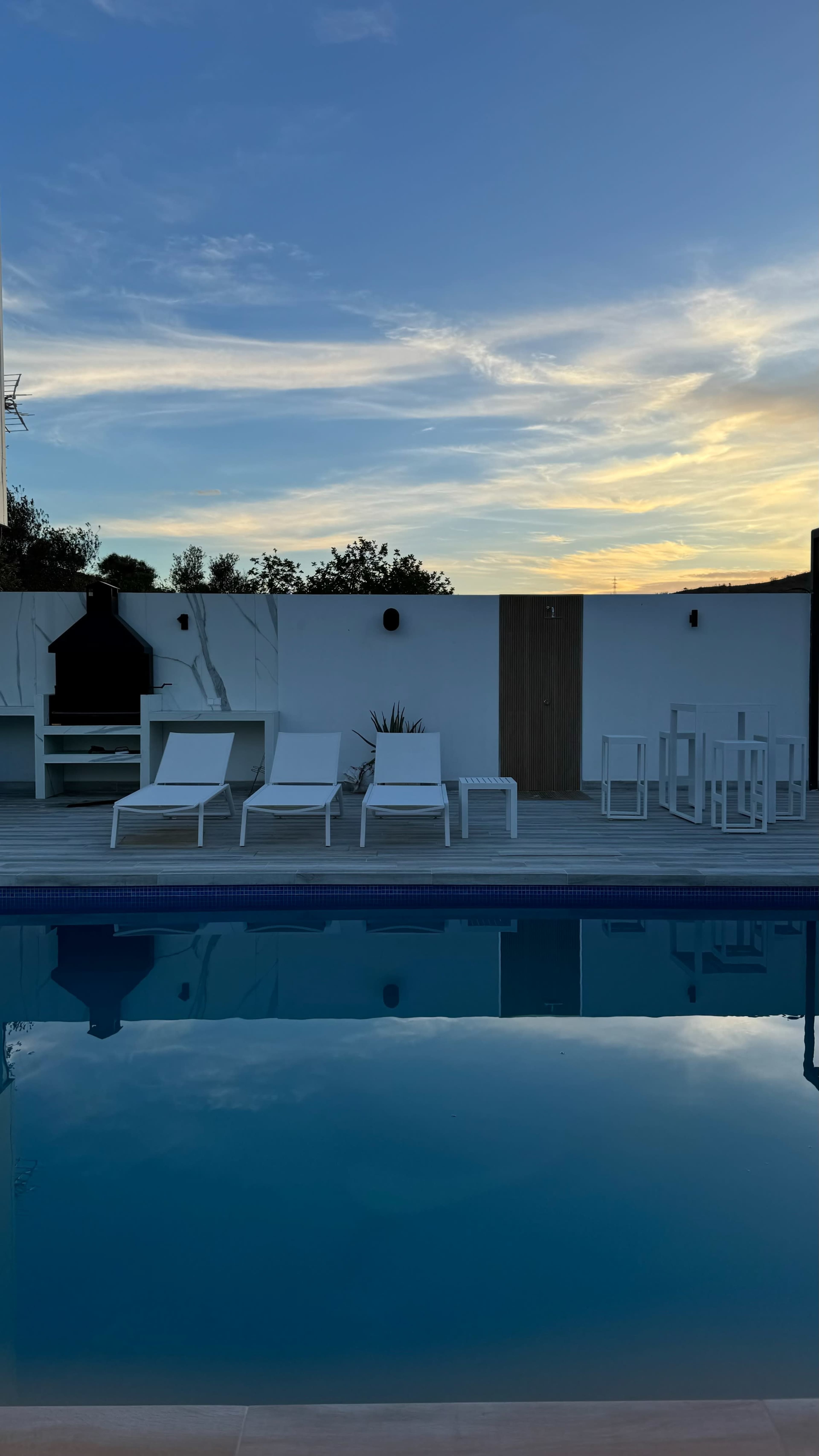 A swimming pool reflects the evening sky, surrounded by white lounge chairs and an outdoor dining area.