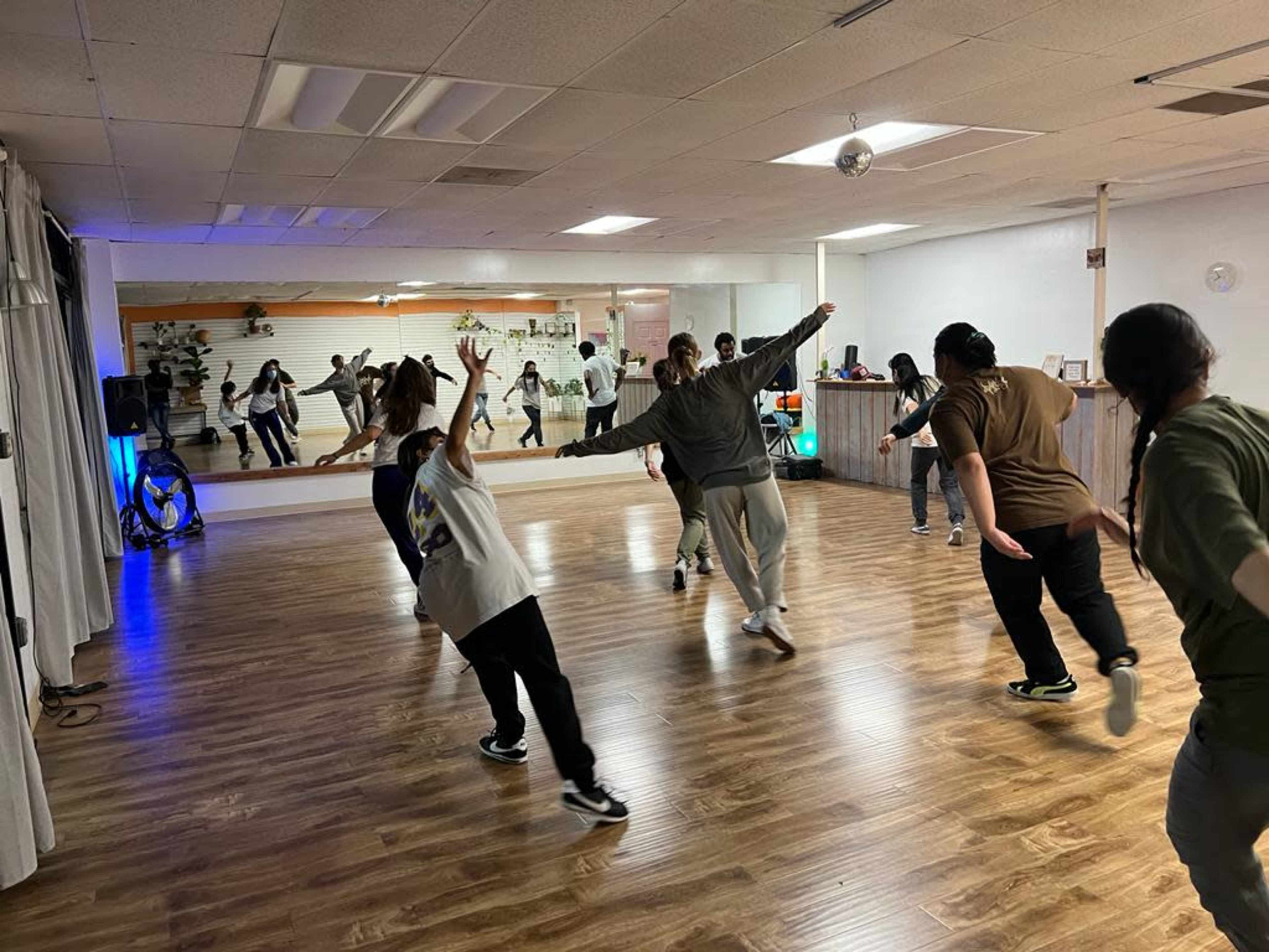 A group of people is dancing in a spacious studio with mirrors and wooden flooring.