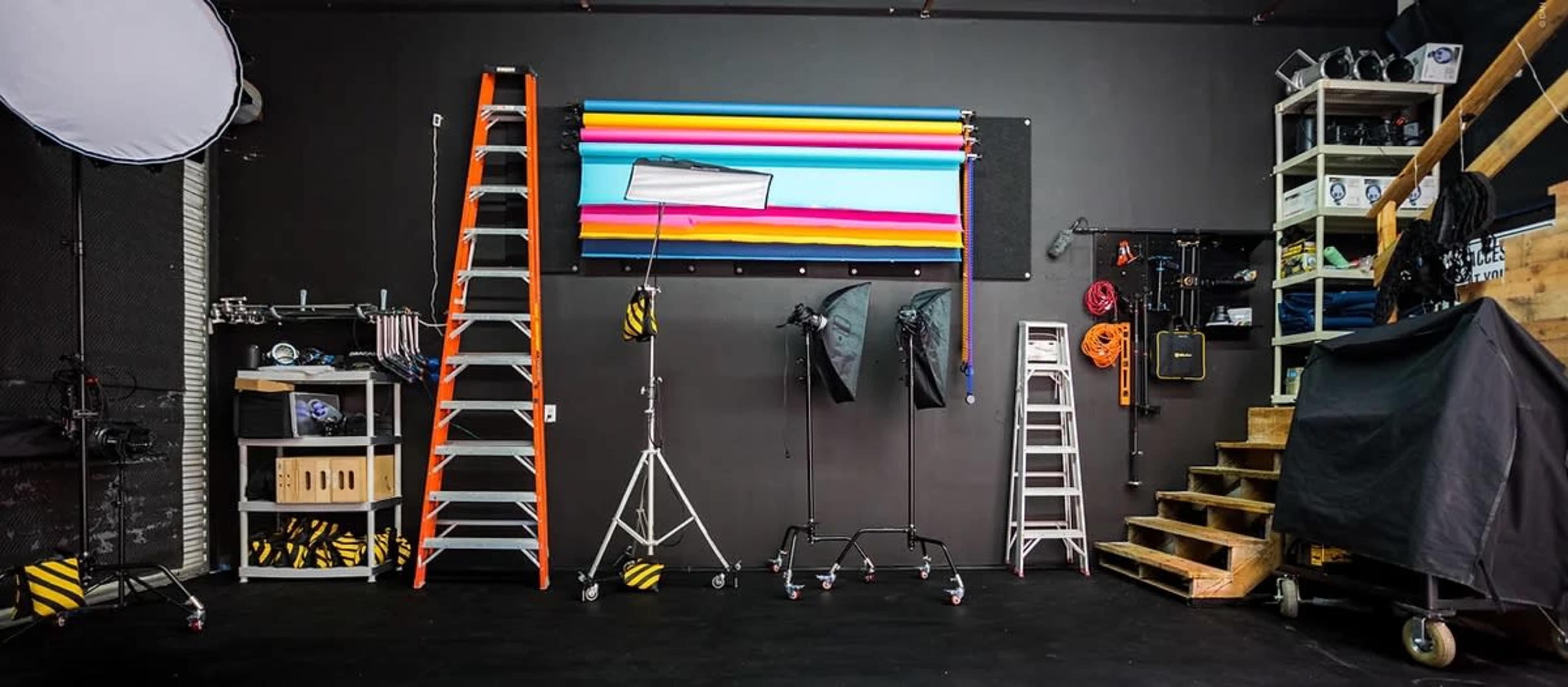 A photography studio equipped with various lighting setups, ladders, and storage shelves against a textured black wall.