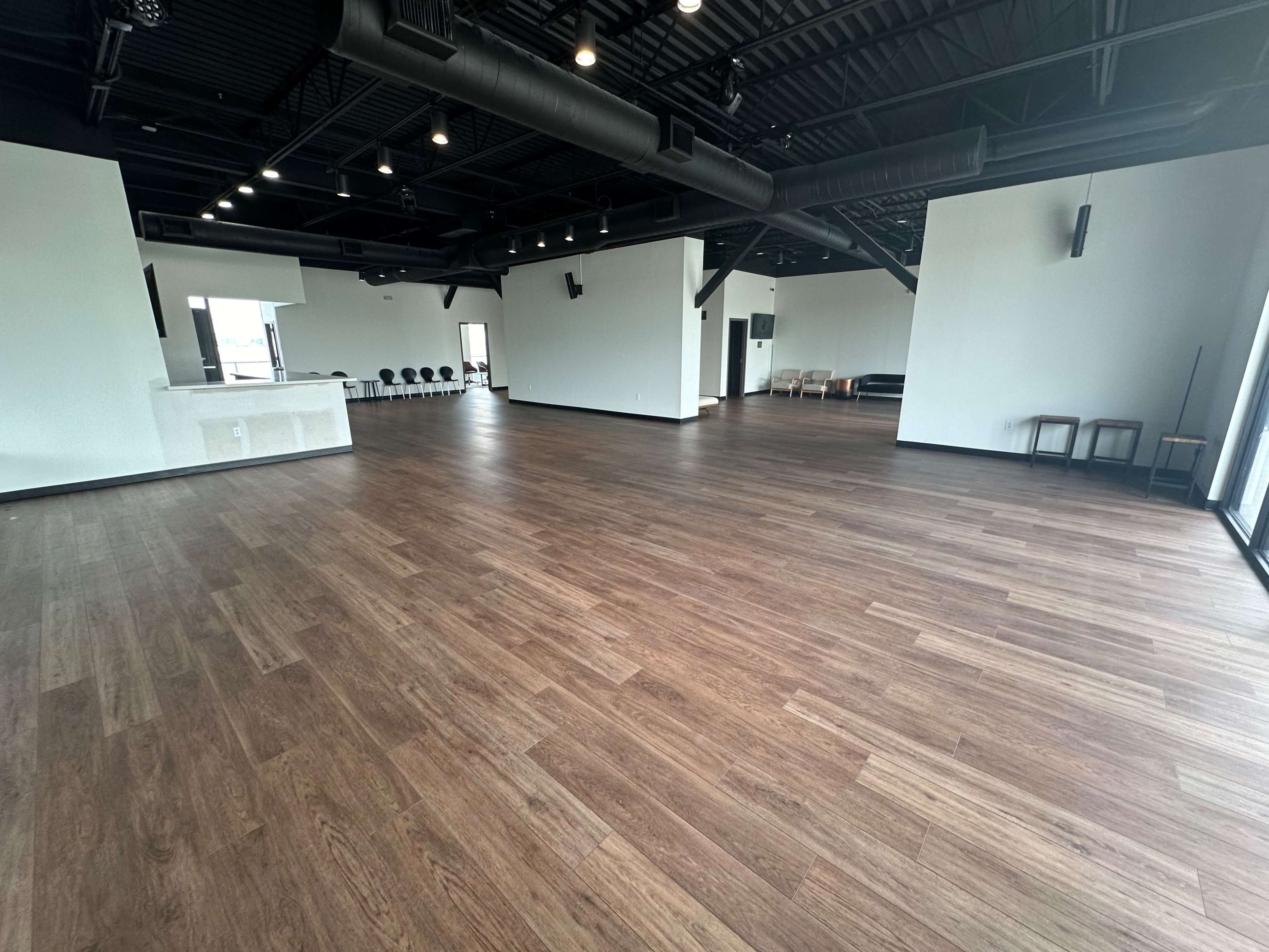 The image shows an empty indoor space with wooden flooring and minimalist design, including a bar area and seating arrangements along the walls.