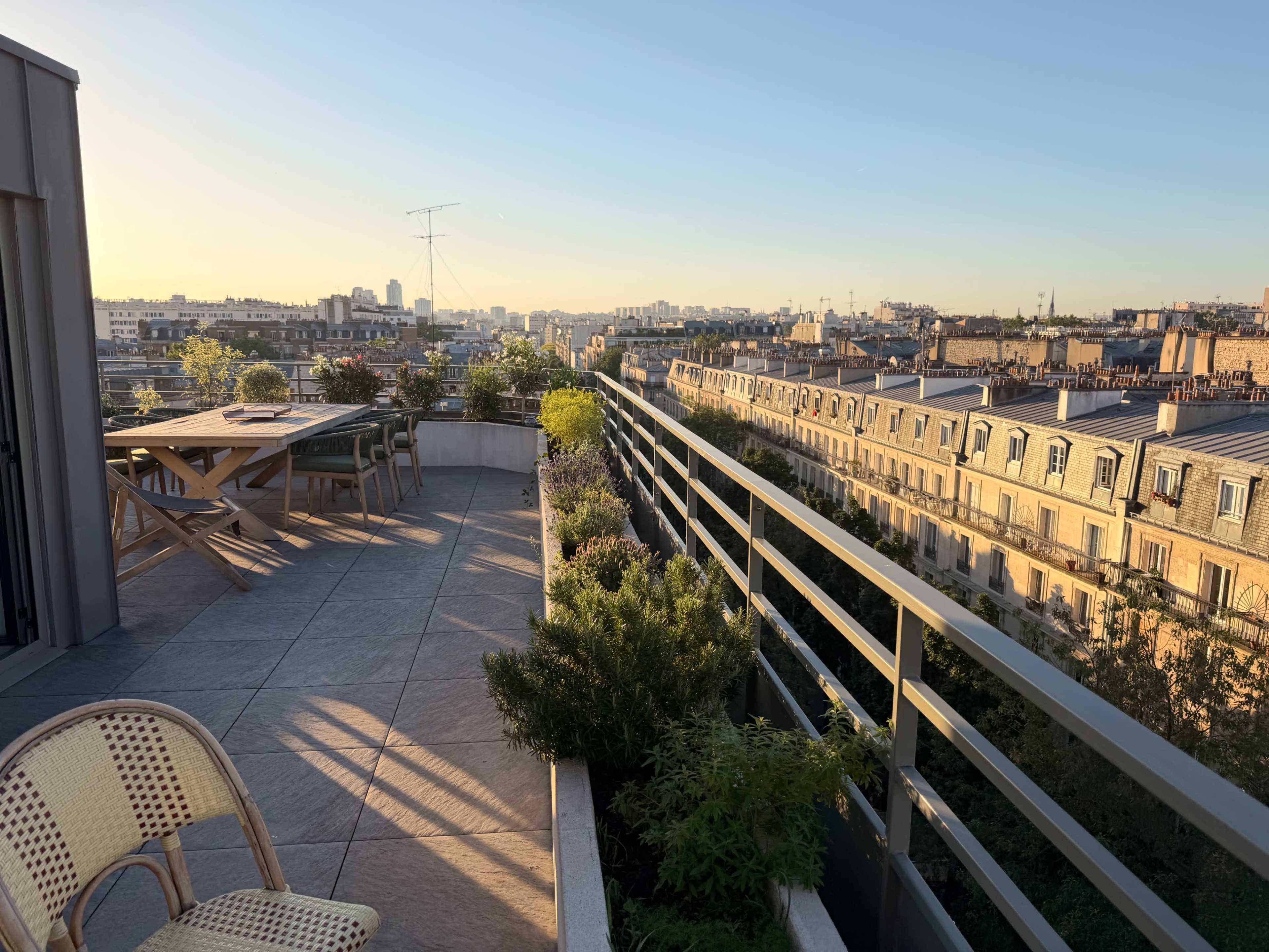 The image shows a rooftop terrace in a city, featuring a wooden table, several planters, and a view of buildings under a clear sky at sunset.