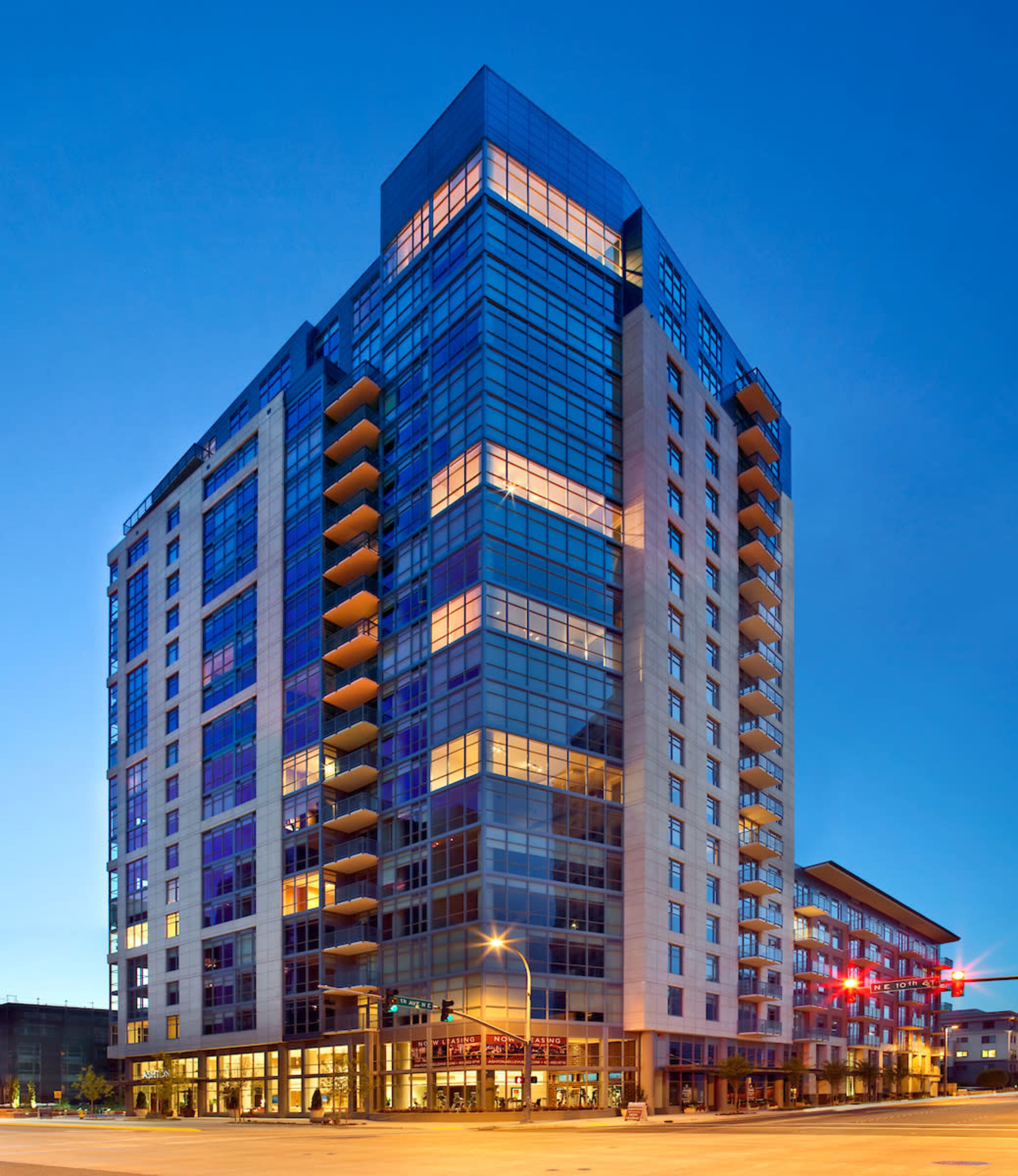 A modern high-rise building with extensive glass windows is located at a street corner, illuminated by dusk light.