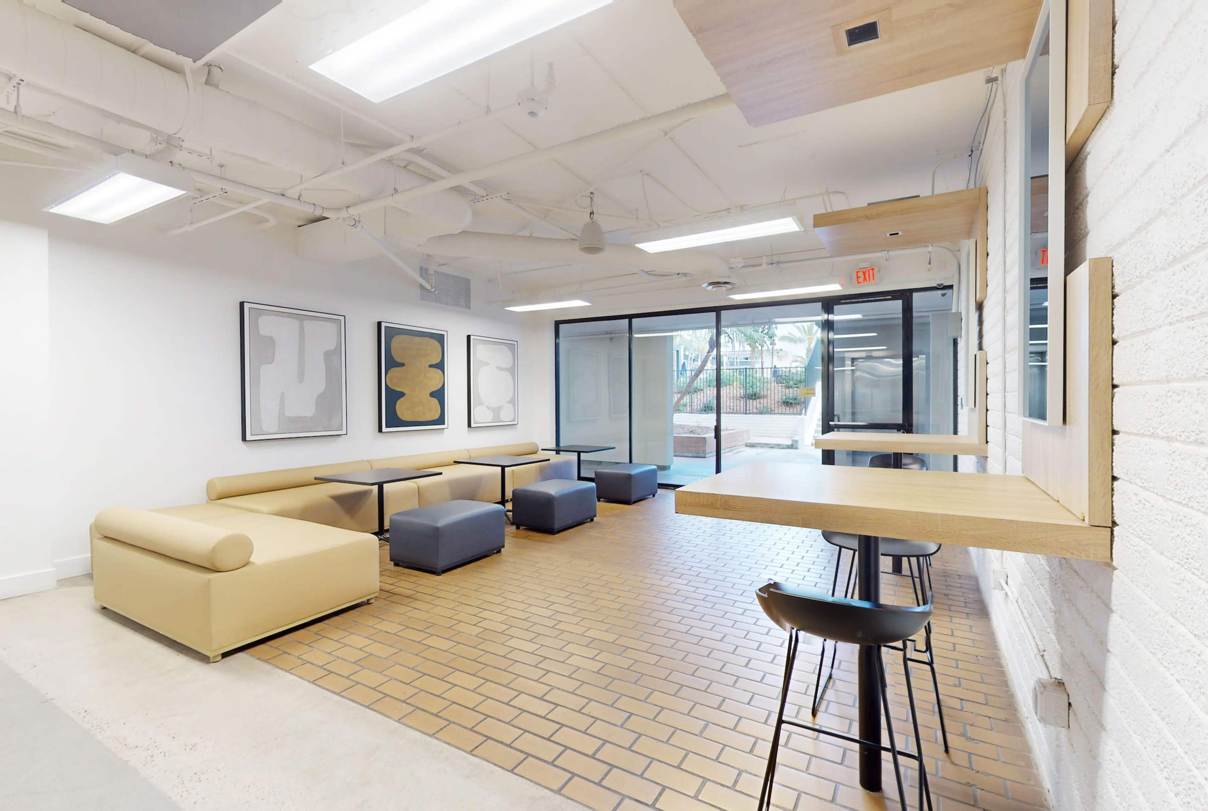 A modern lounge area with beige couches, dark blue ottomans, framed artworks on the walls, and a windowed entrance leading to an outdoor space.