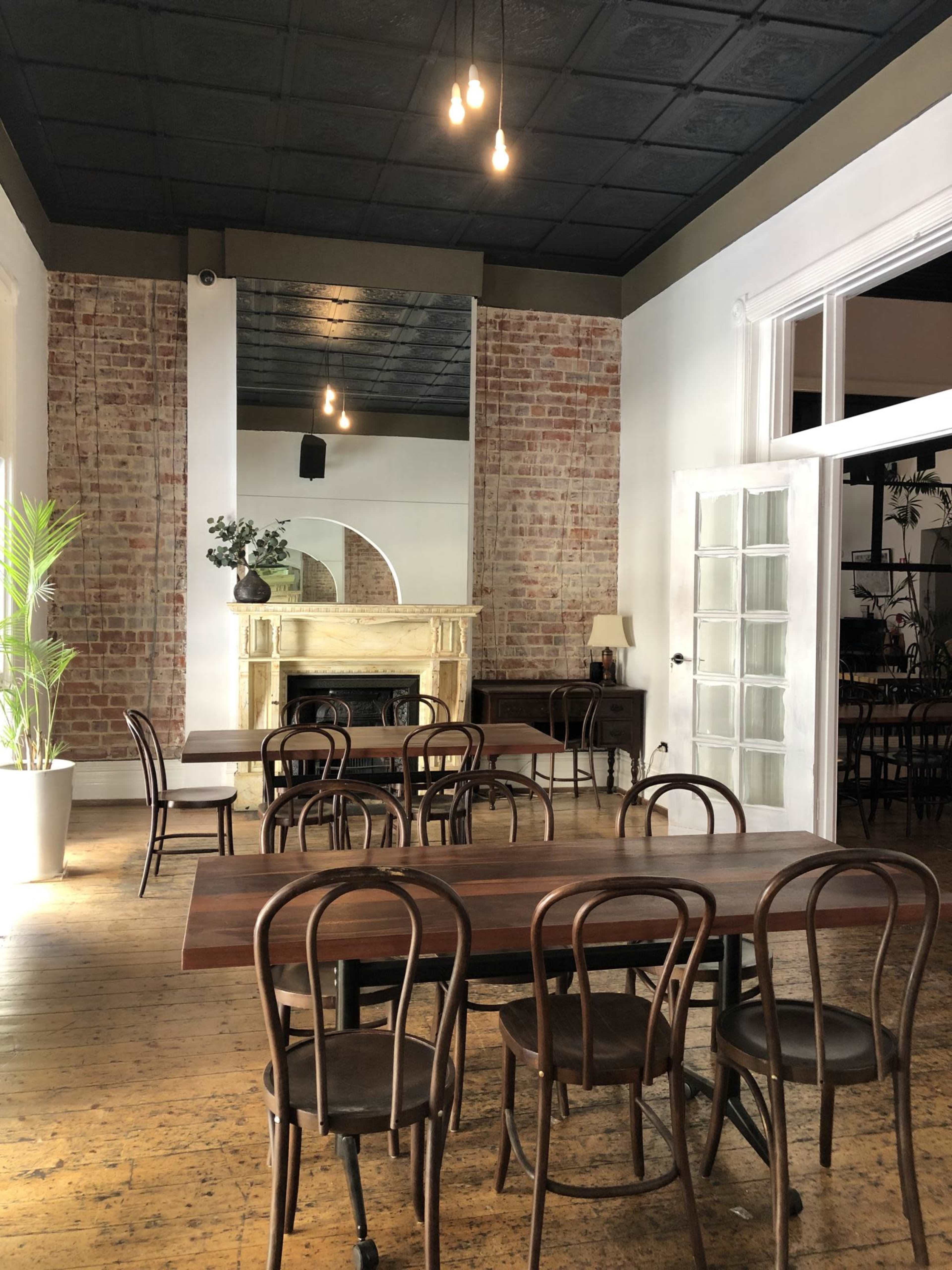A spacious interior of a dining area featuring wooden tables, bentwood chairs, exposed brick walls, and a decorative fireplace.