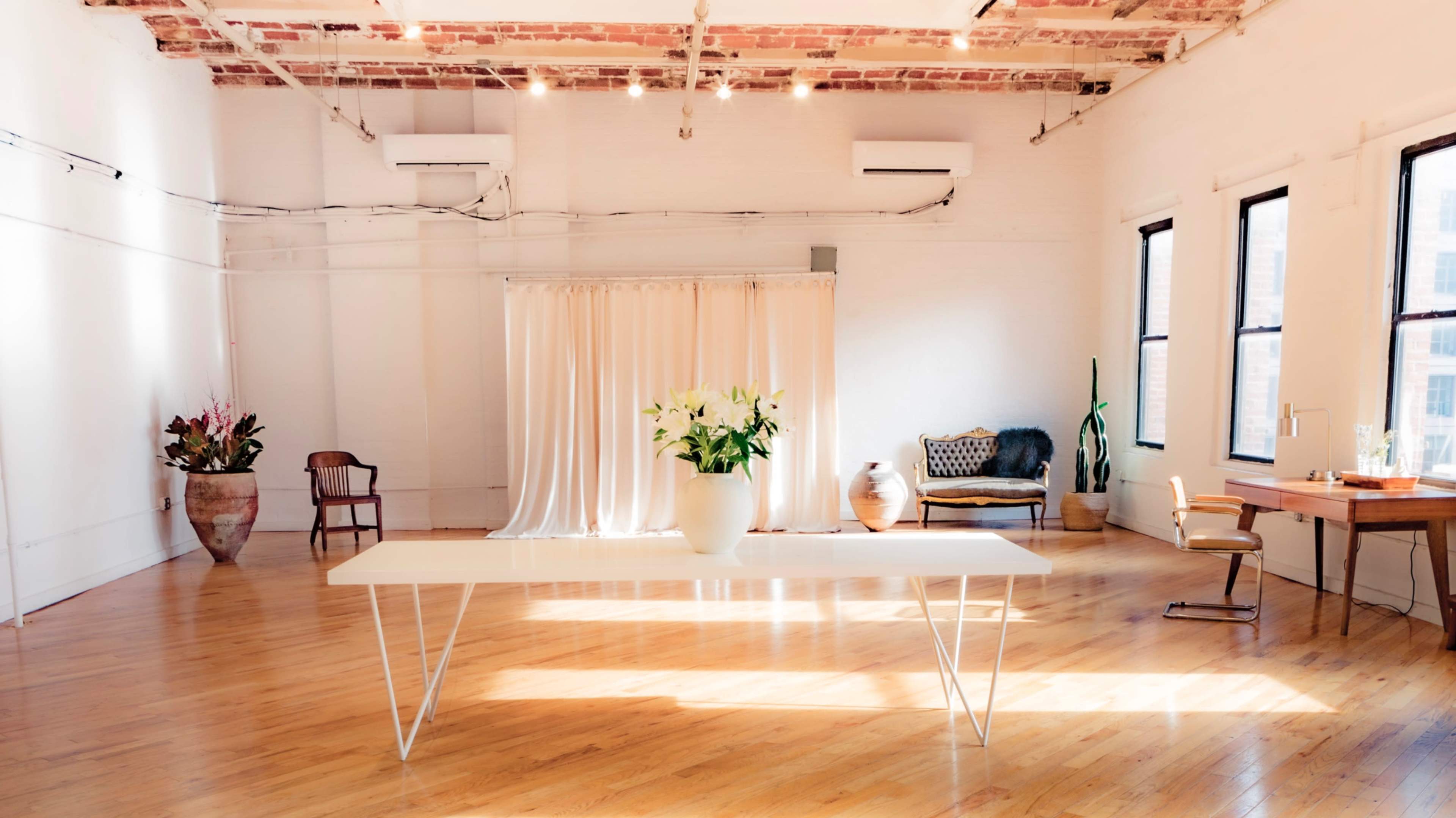 The image shows a spacious, well-lit room featuring a white table with a vase of flowers, a chair, and a small desk against a backdrop of soft curtains and large windows.