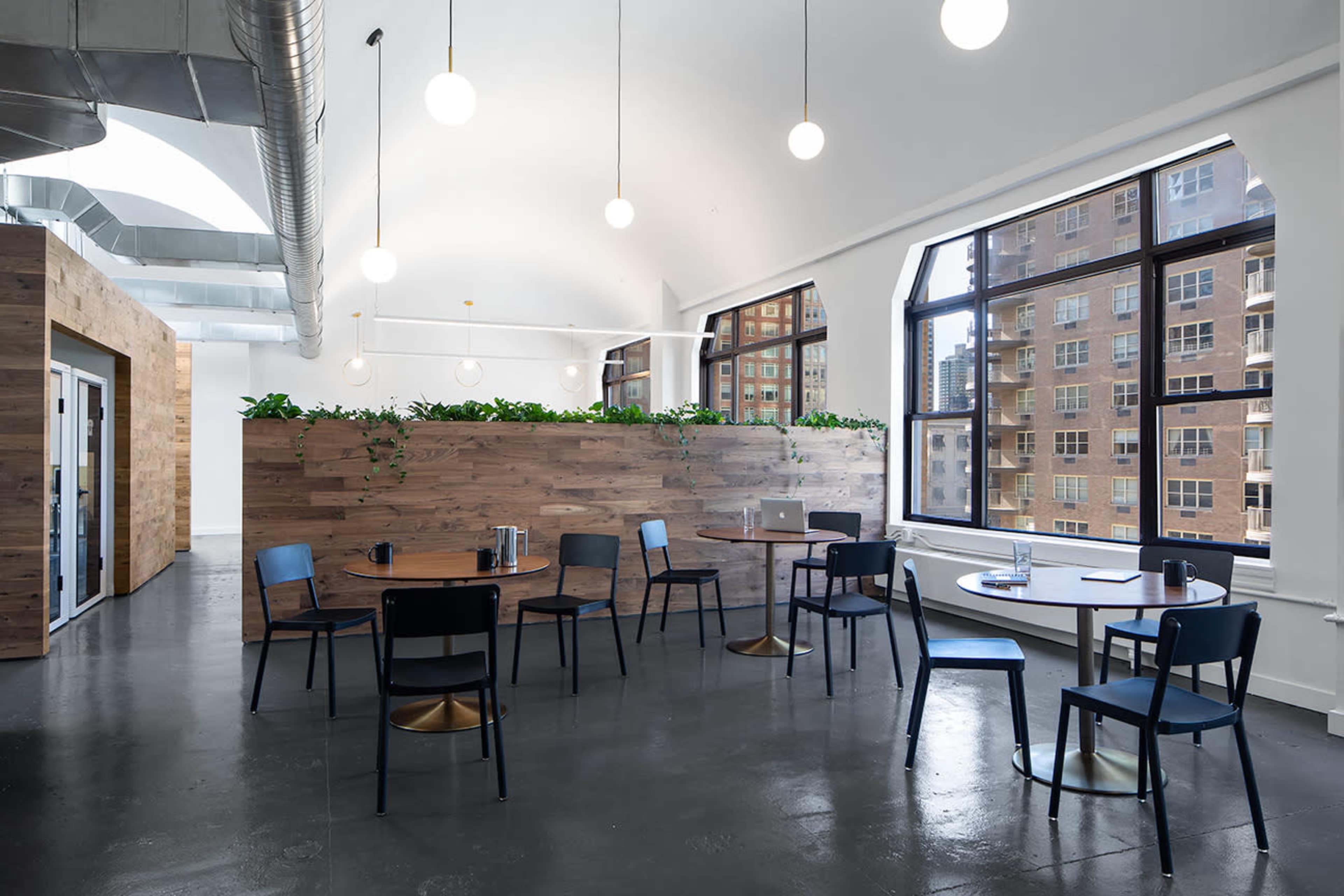 A modern office space with wooden partitioning, several round tables, and chairs, illuminated by pendant lights.