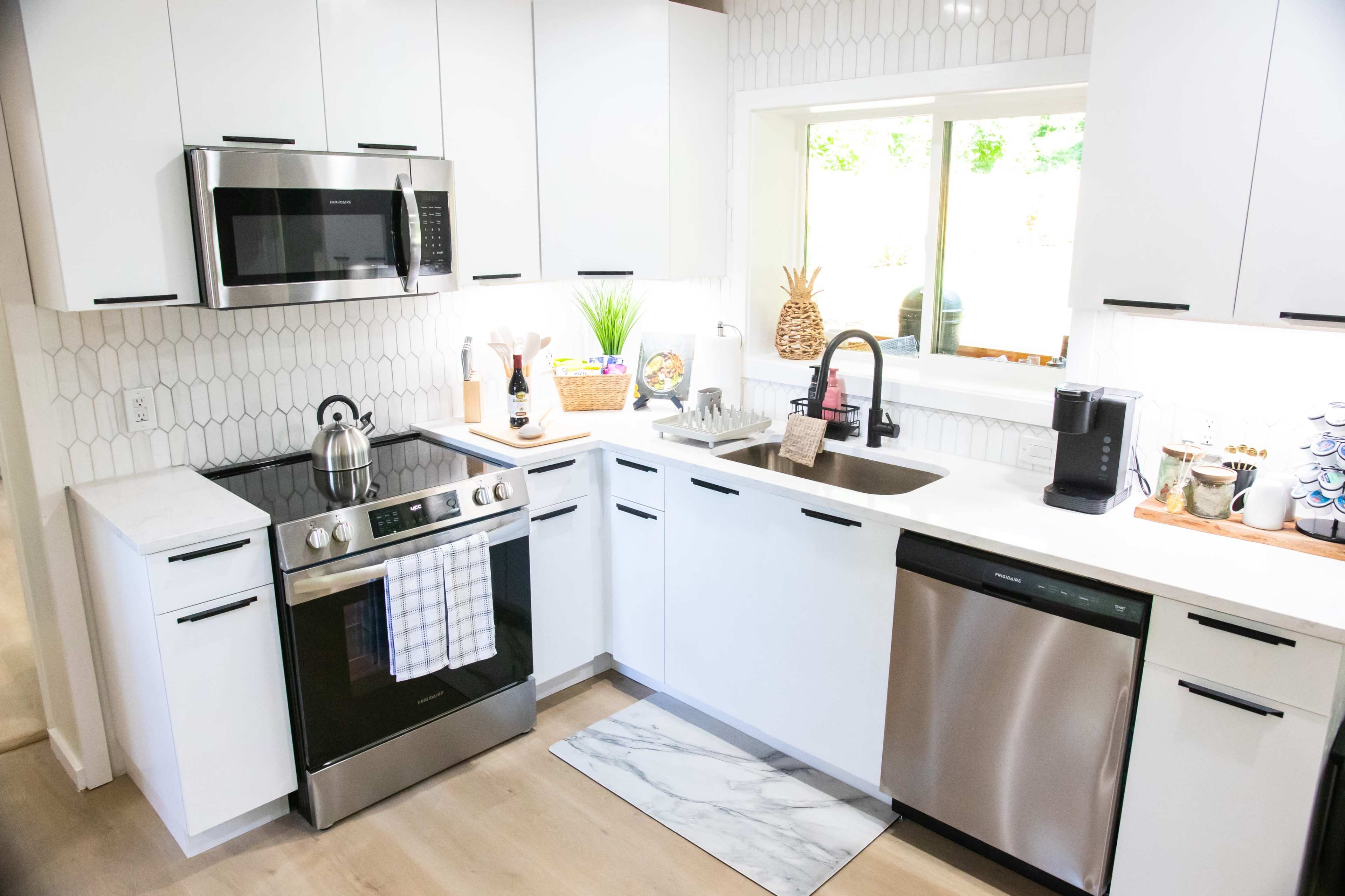 A modern kitchen with white cabinetry, a stainless steel oven and microwave, a black faucet, and a window above the sink that lets in natural light.