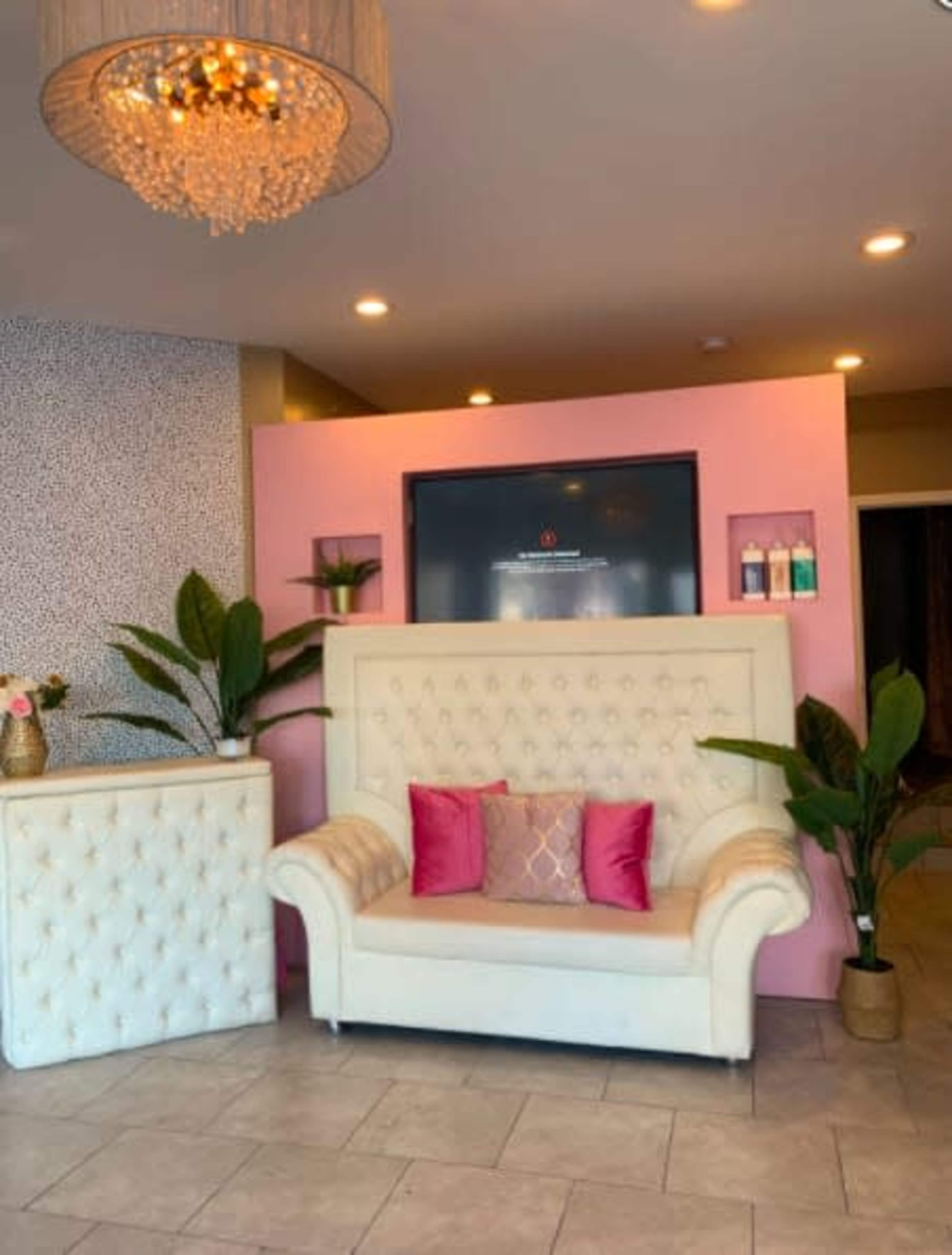 A modern reception area featuring a white tufted sofa with pink cushions, a decorative chandelier, and potted plants against a pink and patterned wall.