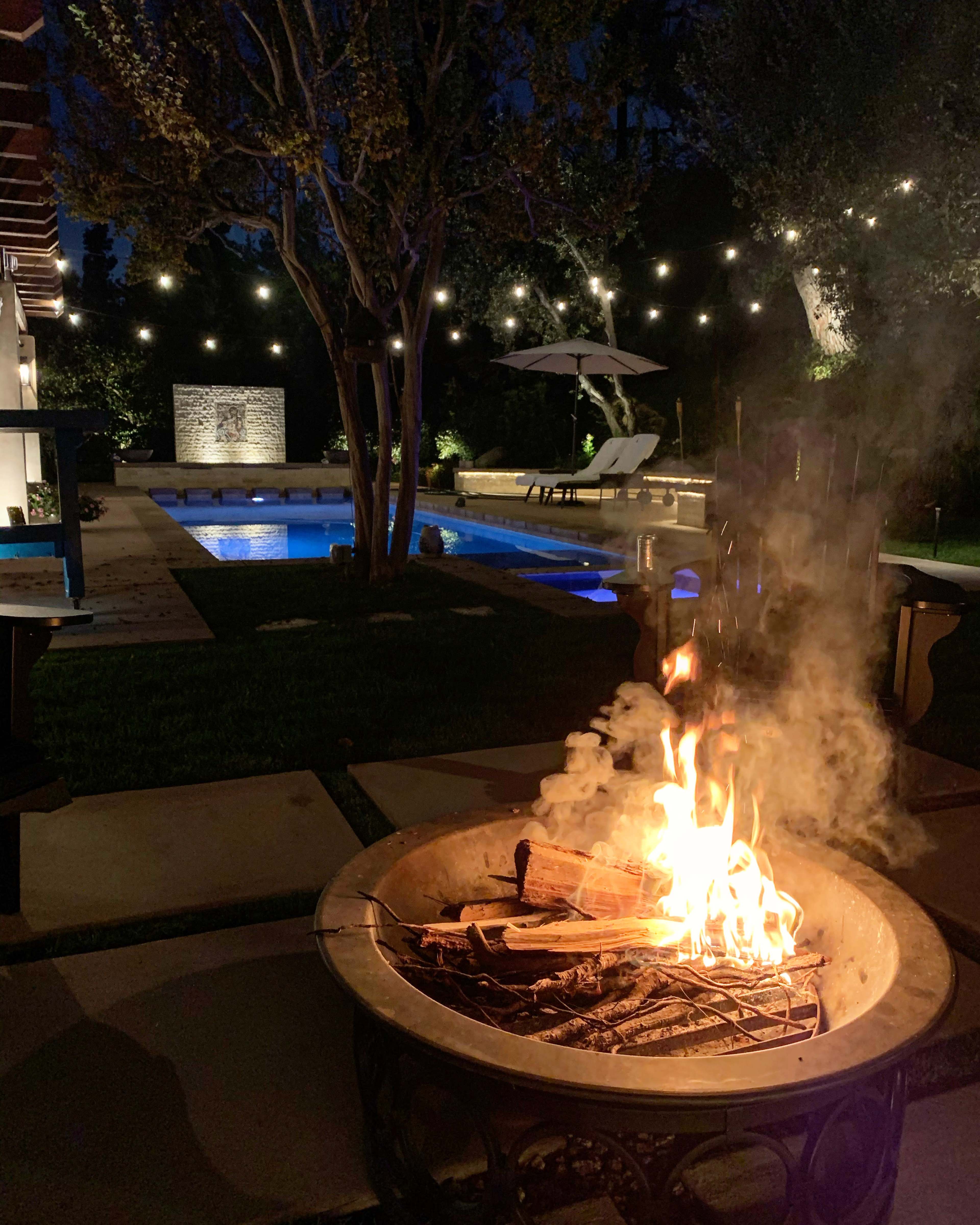 A backyard scene at night features a fire pit with flames, surrounded by stone pathways, a swimming pool, and softly lit trees.