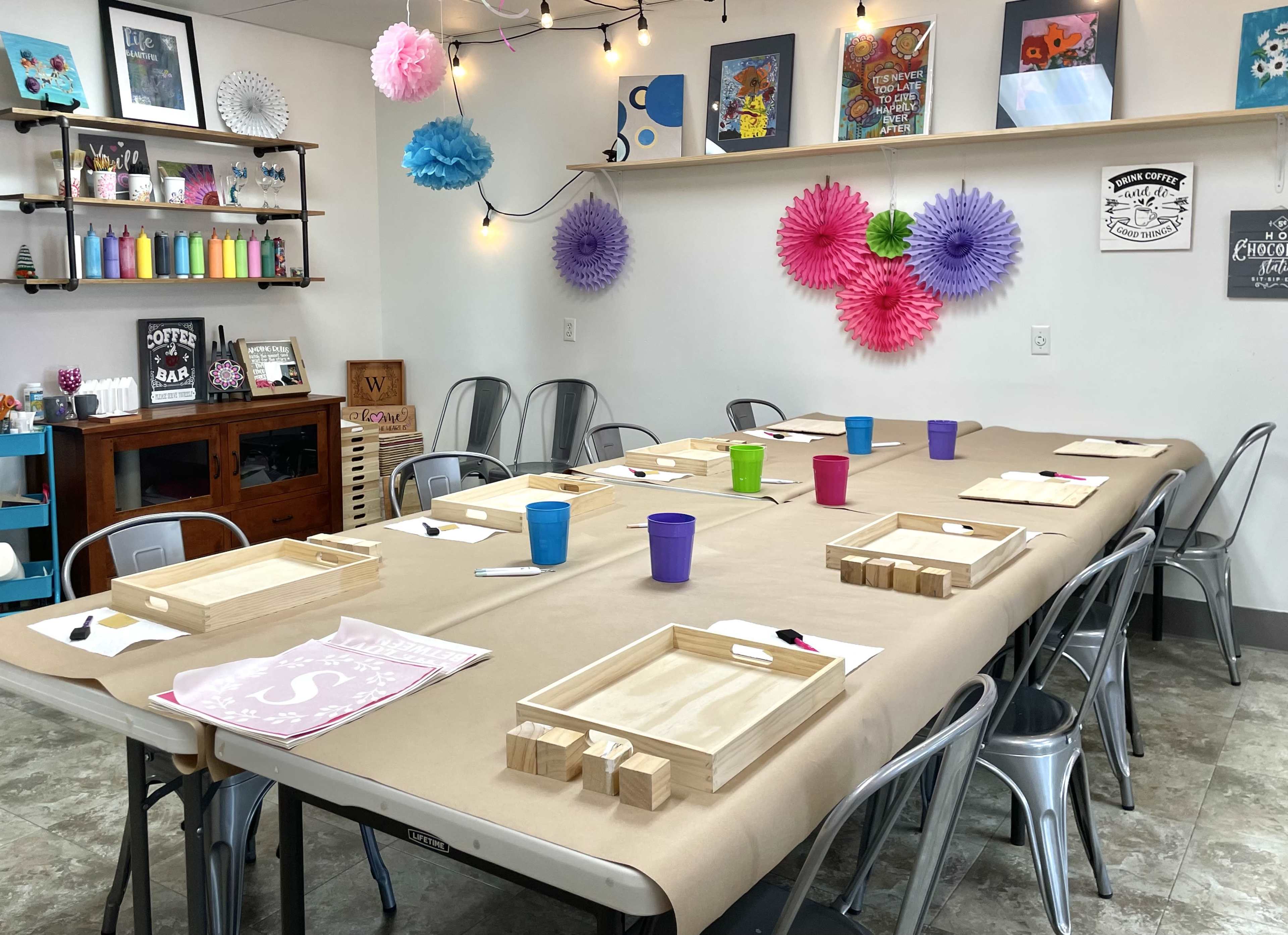 A large, rectangular table is set for an event, covered with brown paper, and surrounded by metal chairs, with colorful cups and wooden trays arranged neatly at each place, and decorations hanging on the walls.