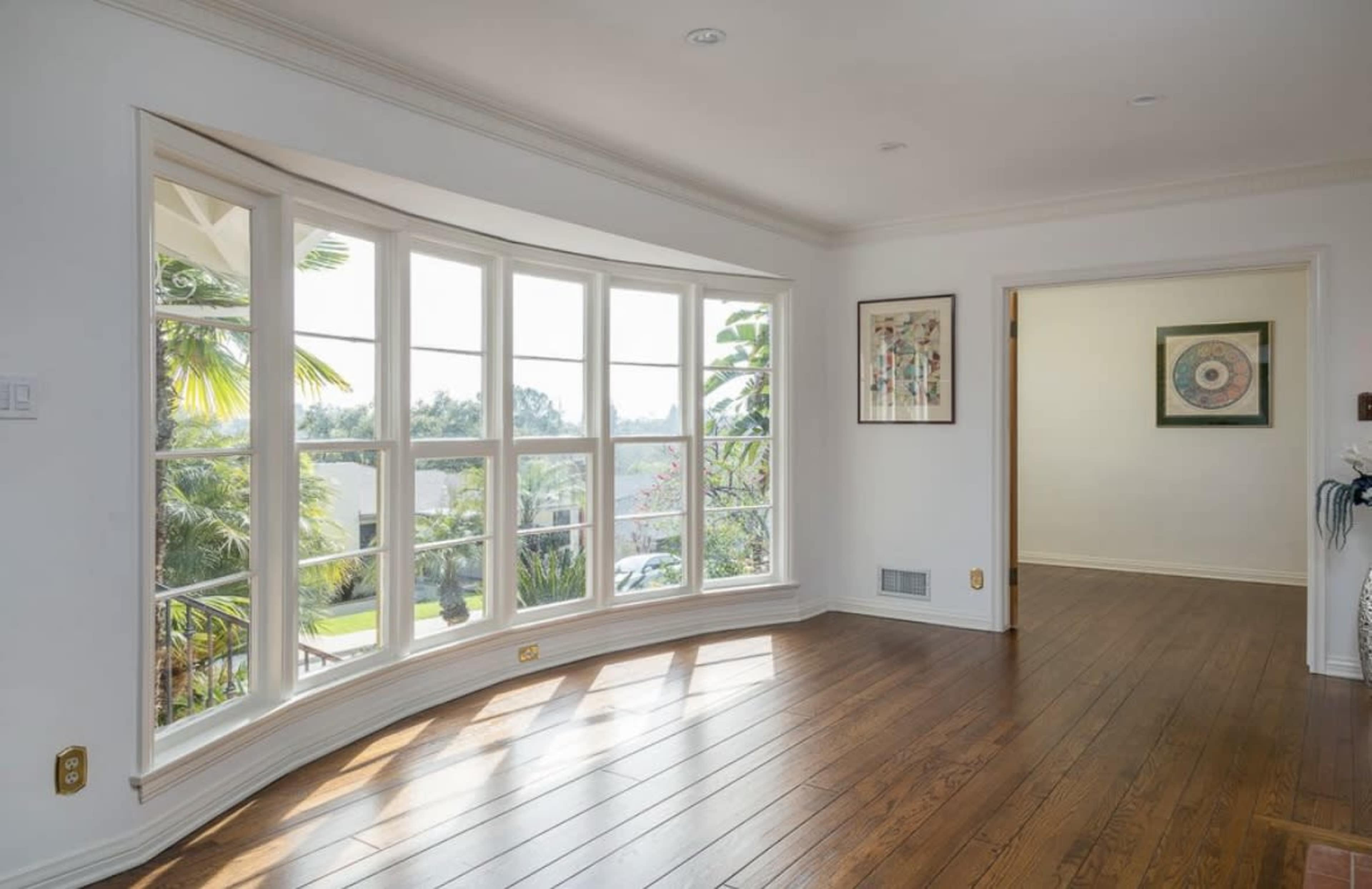 The image shows a bright room with large bay windows facing a garden, featuring wooden flooring and adjacent to another room.