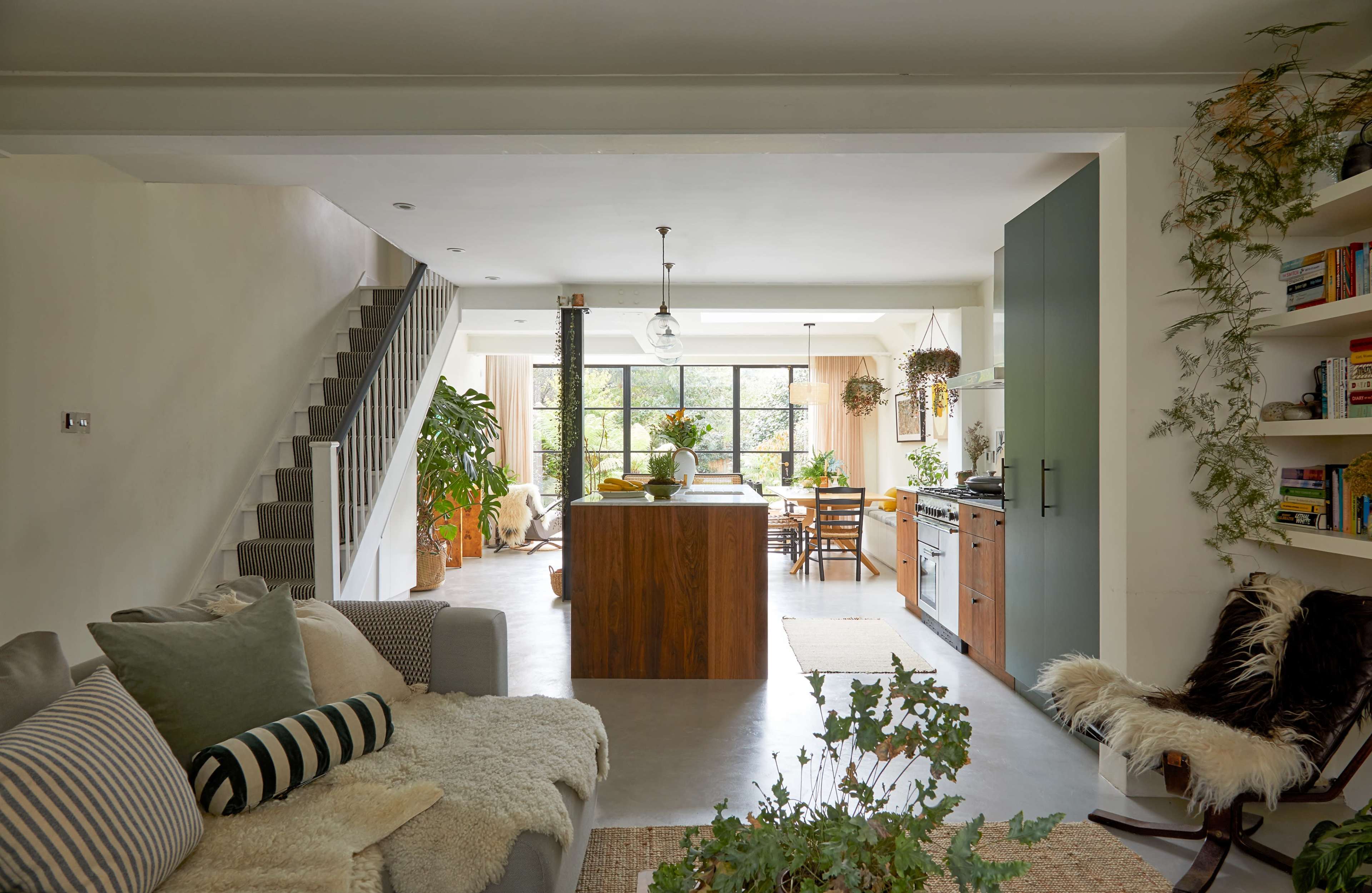 The image shows a modern open-concept kitchen and living area, featuring a staircase, a large window, and plants throughout the space.