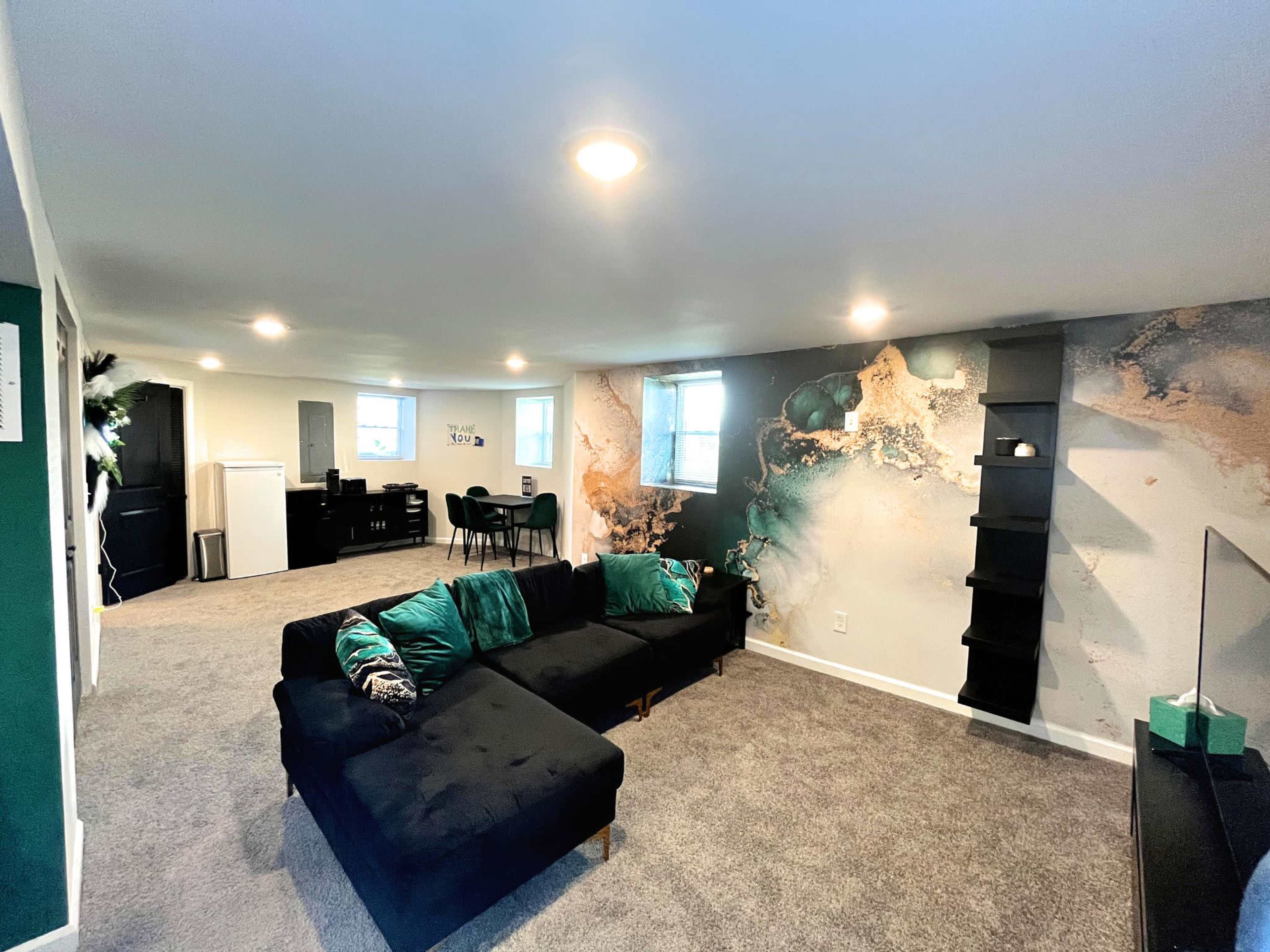 A spacious living area featuring a black sectional sofa, a decorative wall with a multi-colored design, and a dining area with a table and chairs in the background.