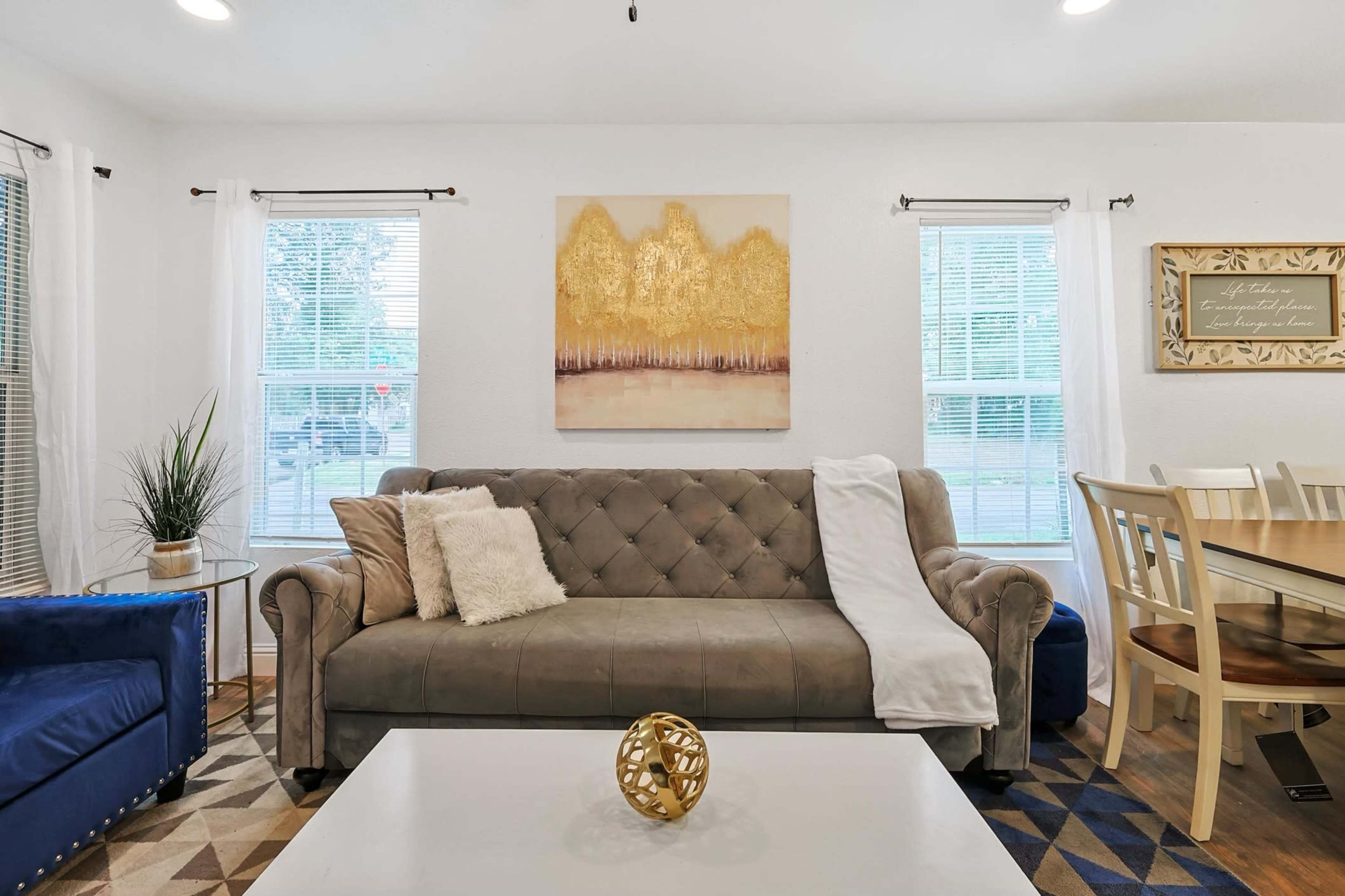 A living room with a gray sofa, a white coffee table, and a dining area nearby, featuring large windows and a painting above the sofa.