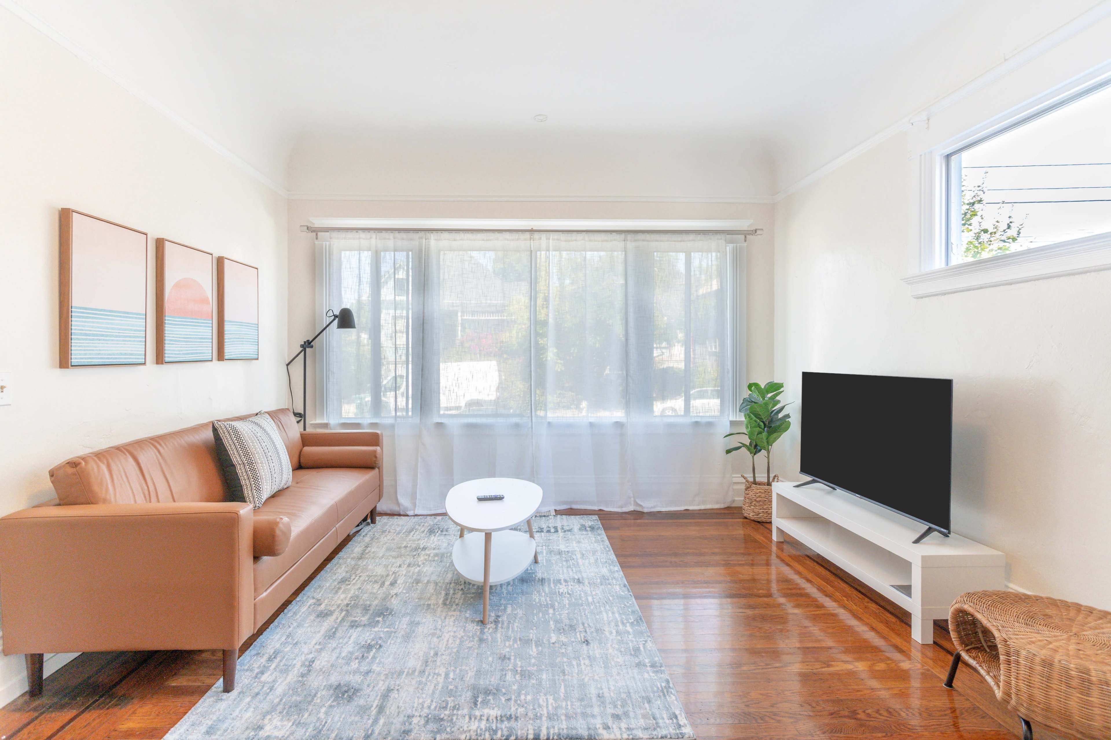 A modern living room features a brown leather sofa, a round coffee table, a TV stand with a flat-screen TV, and large windows draped with sheer curtains.