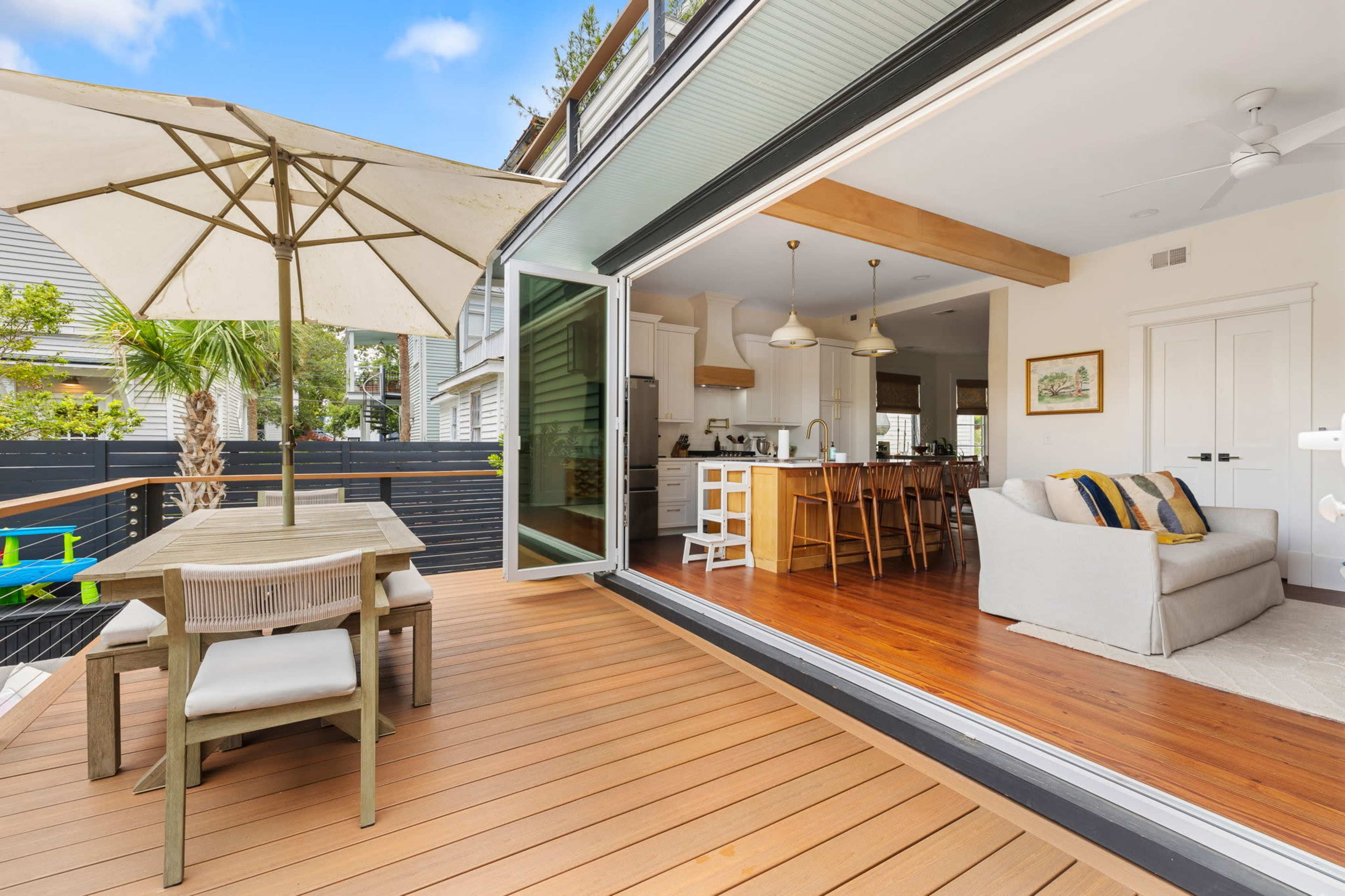 A wooden deck features a dining table with an umbrella and opens to an interior space with a kitchen and living area.