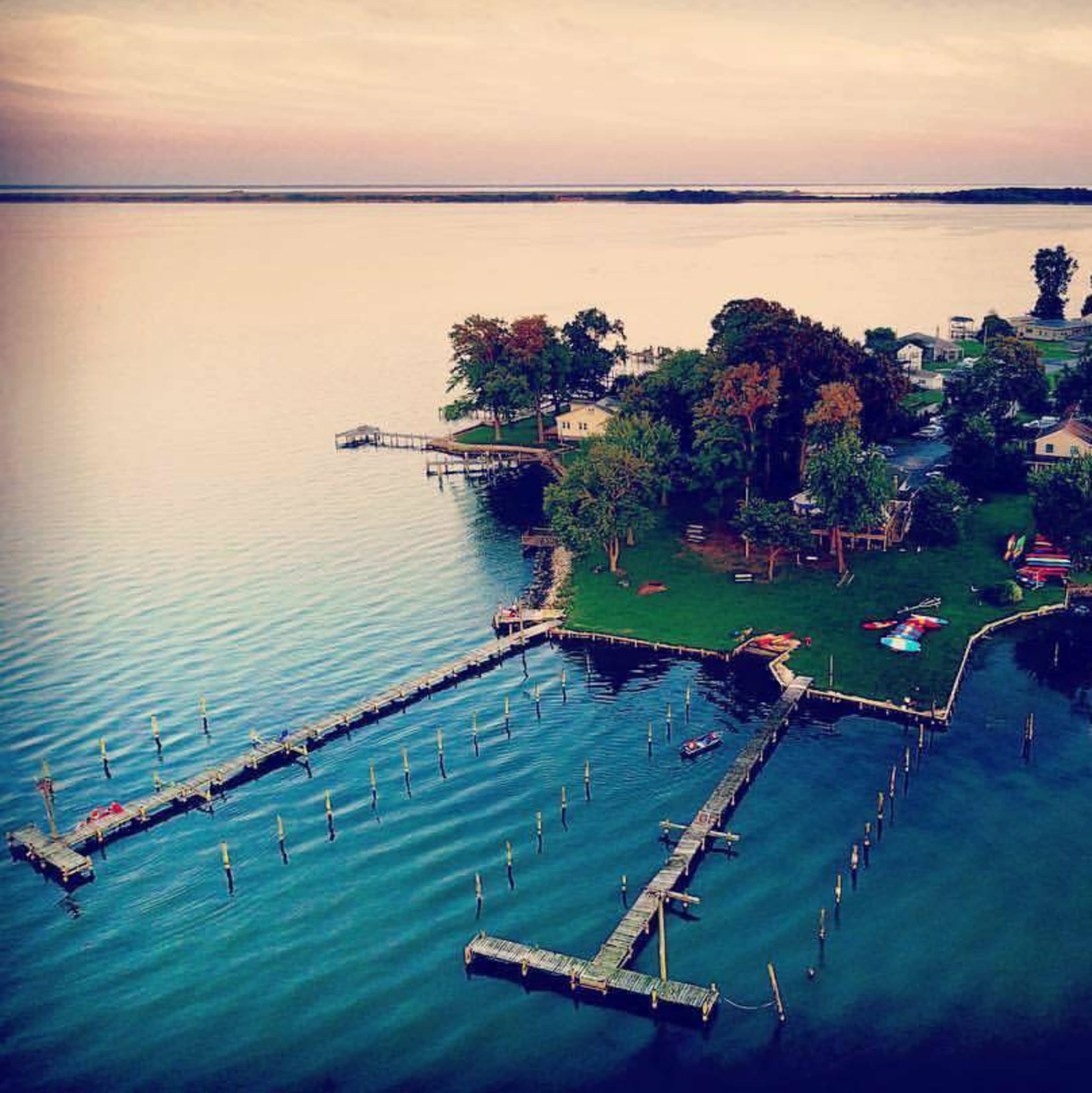 Aerial view of a dock extending into calm waters, surrounded by lush greenery and a few houses on the shoreline.