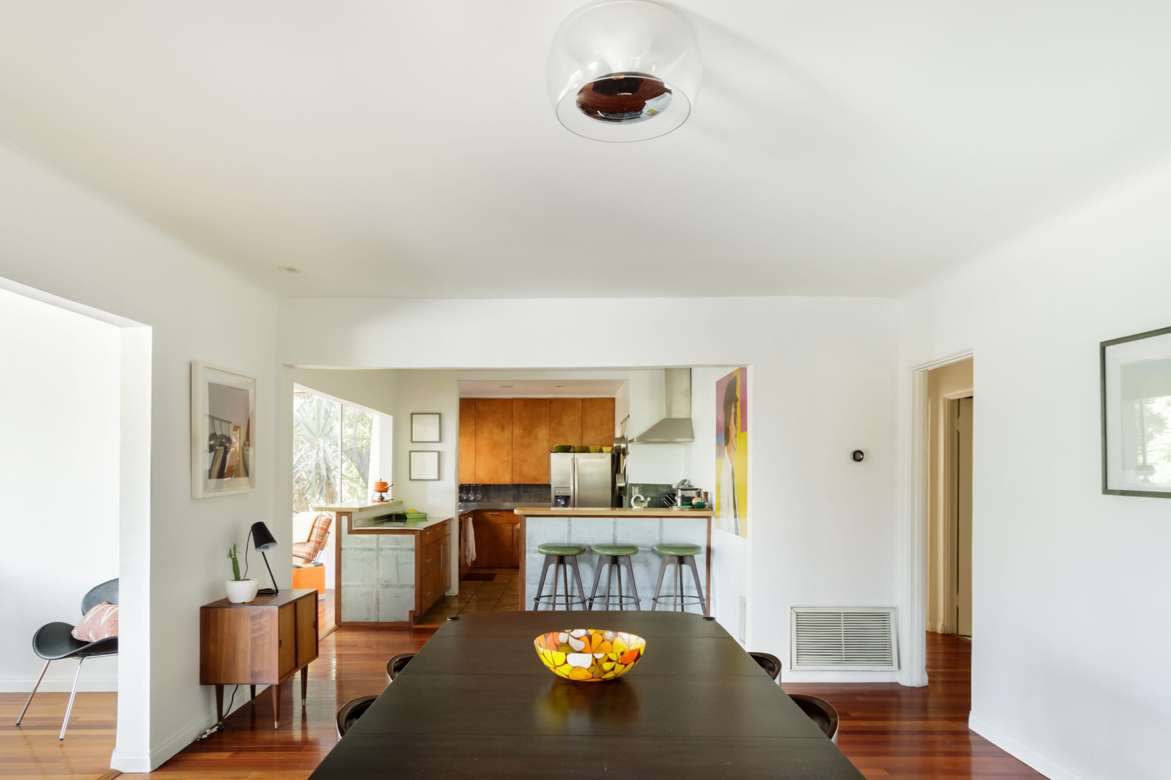 A bright, open dining area connected to a modern kitchen, featuring a large dark wooden table and a colorful fruit bowl as a centerpiece.