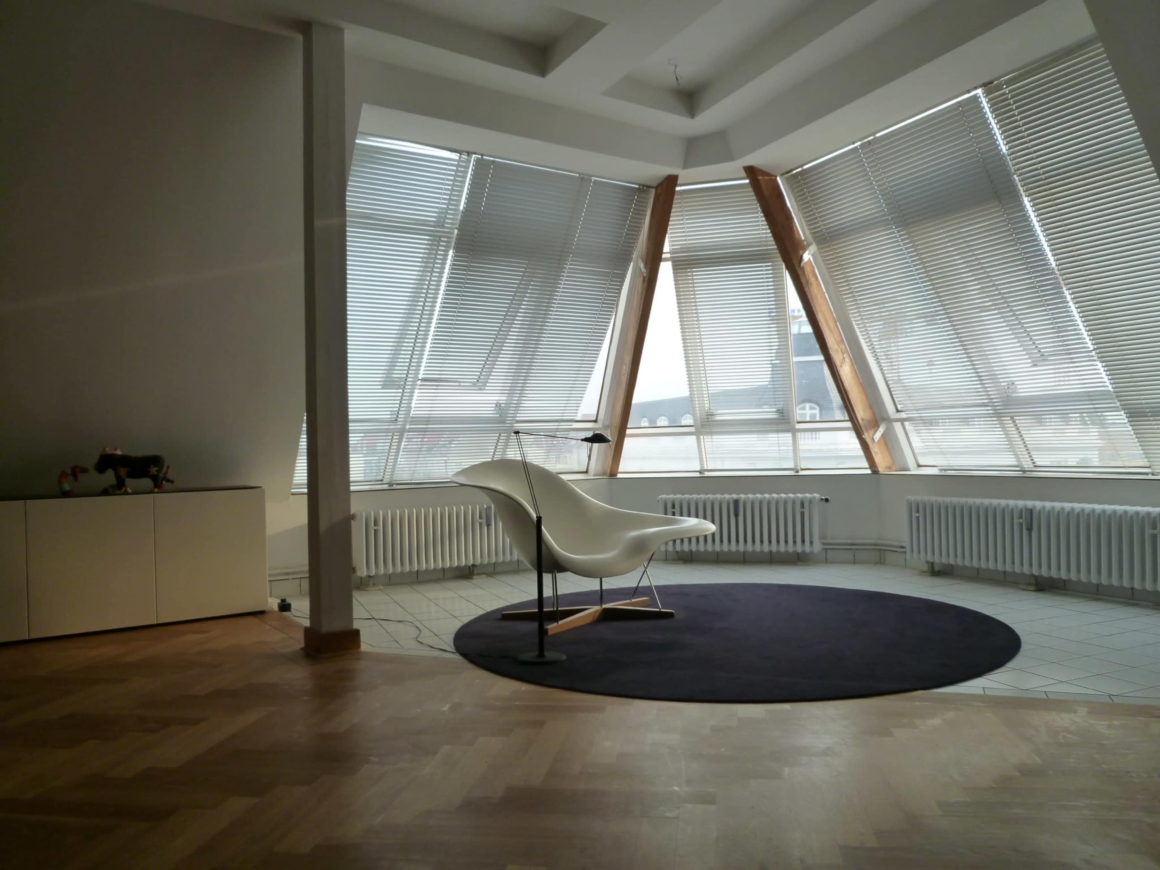 The image shows a spacious modern room with large windows, wooden structural elements, and a minimalist white chair on a circular dark rug.