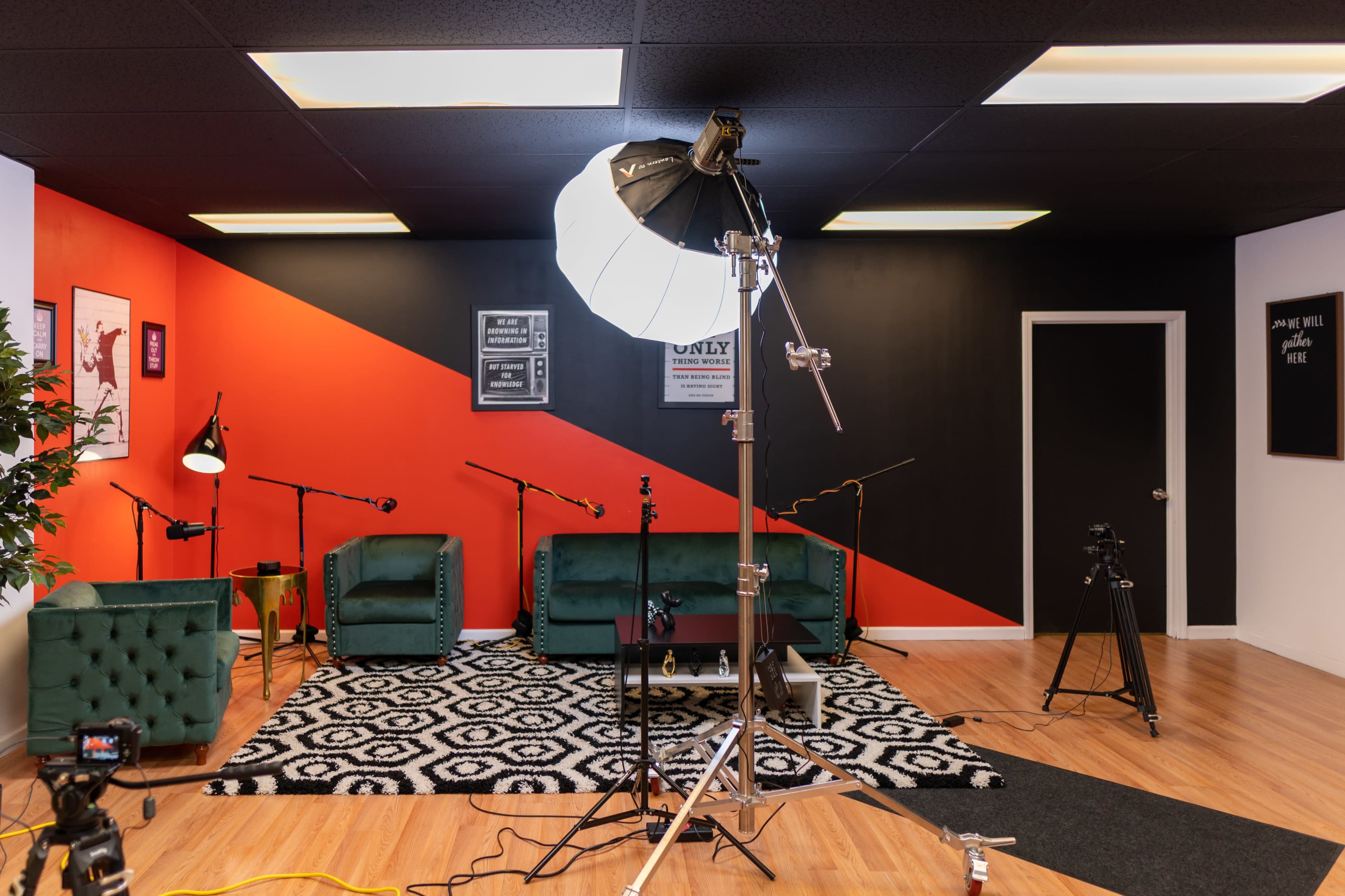 A modern studio setup featuring green velvet chairs, a patterned rug, lighting equipment, and microphones against a backdrop of black and red walls.