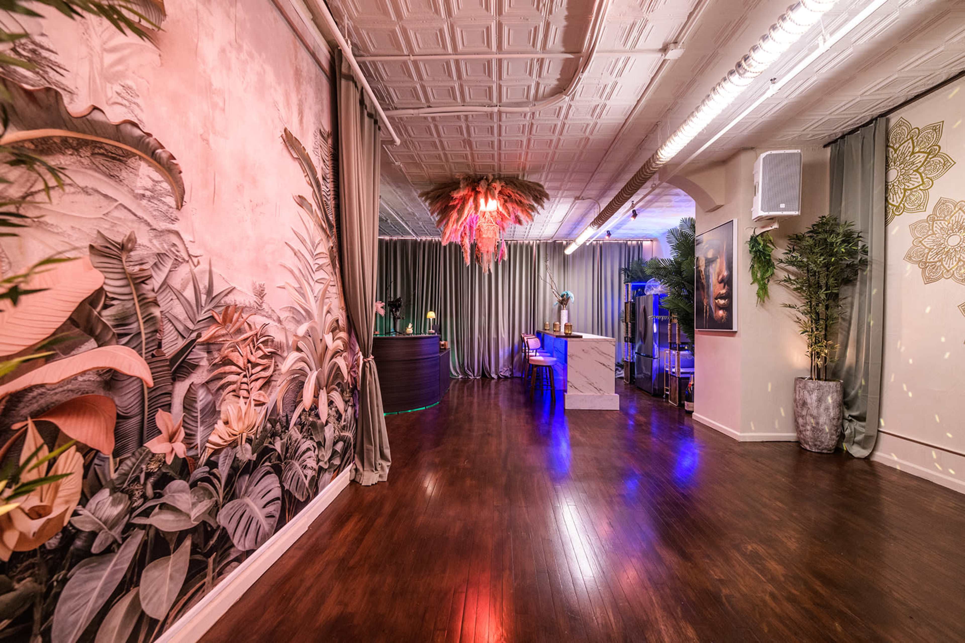 The image shows a stylish interior space with eclectic decor, featuring a tropical mural, a bar area, and soft lighting.