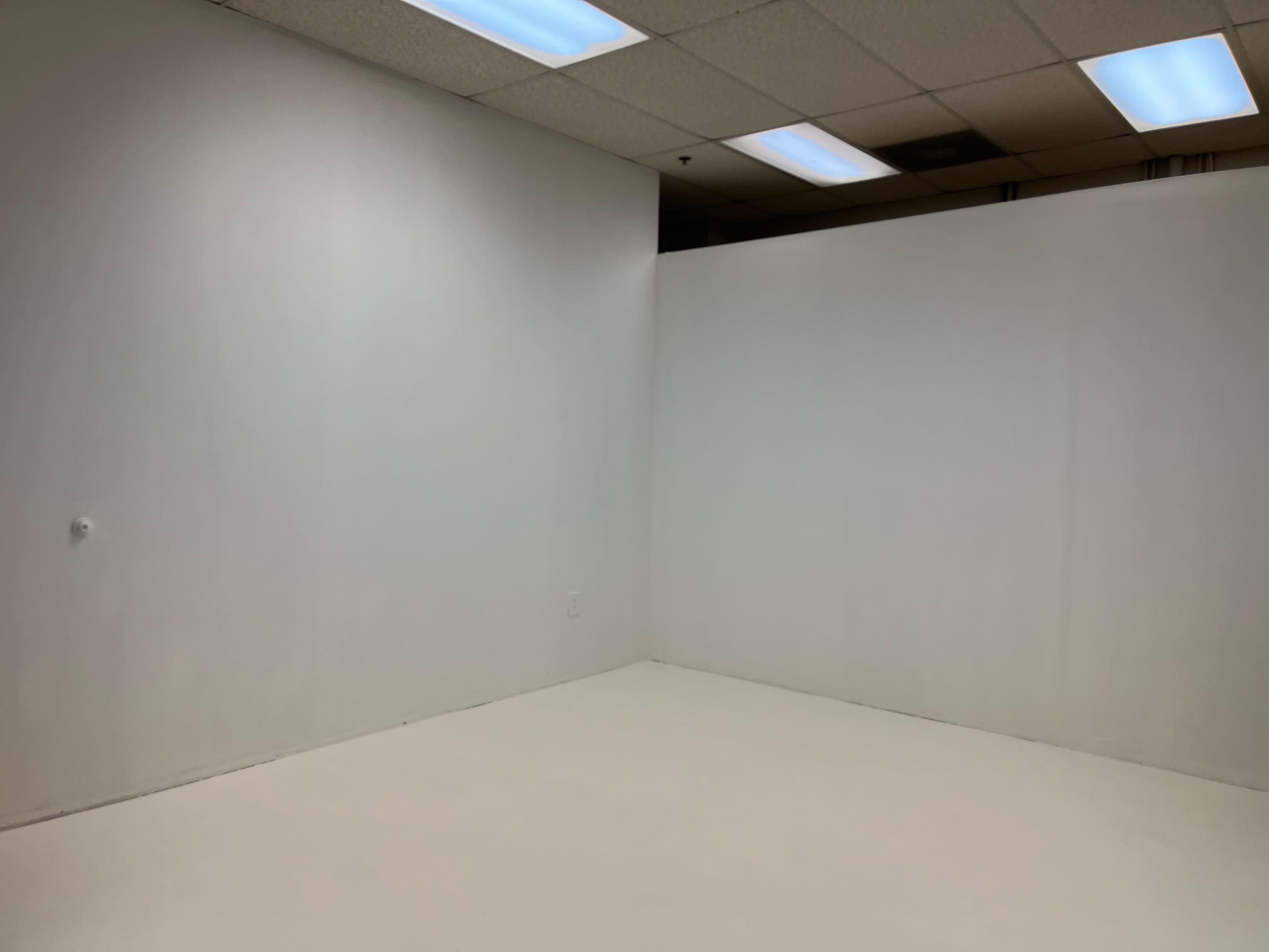 An empty room with white walls and a white floor, featuring a corner and fluorescent ceiling lights.