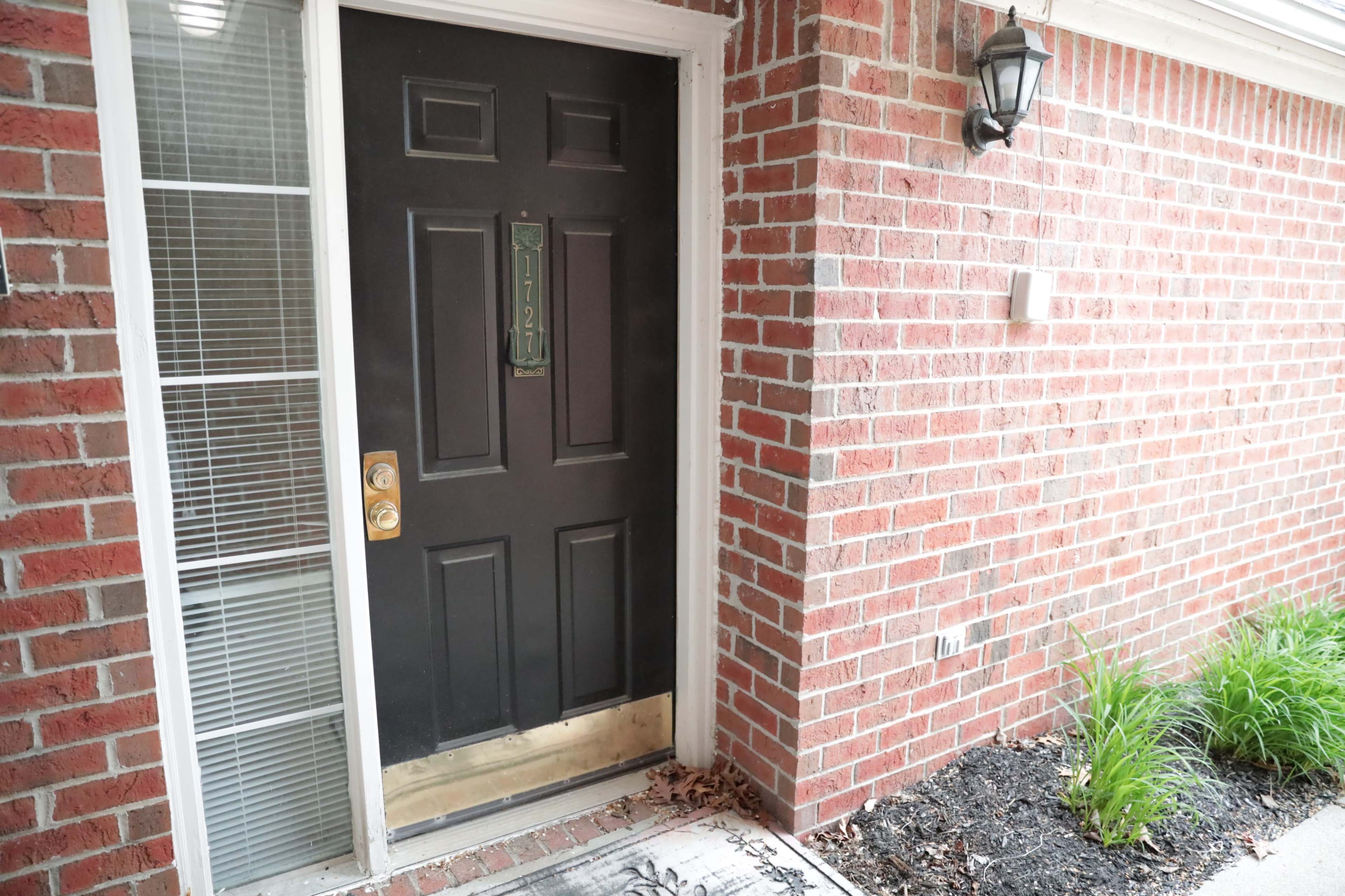 A black front door is framed by brick walls, with a small light fixture and green plants beside a welcome mat.
