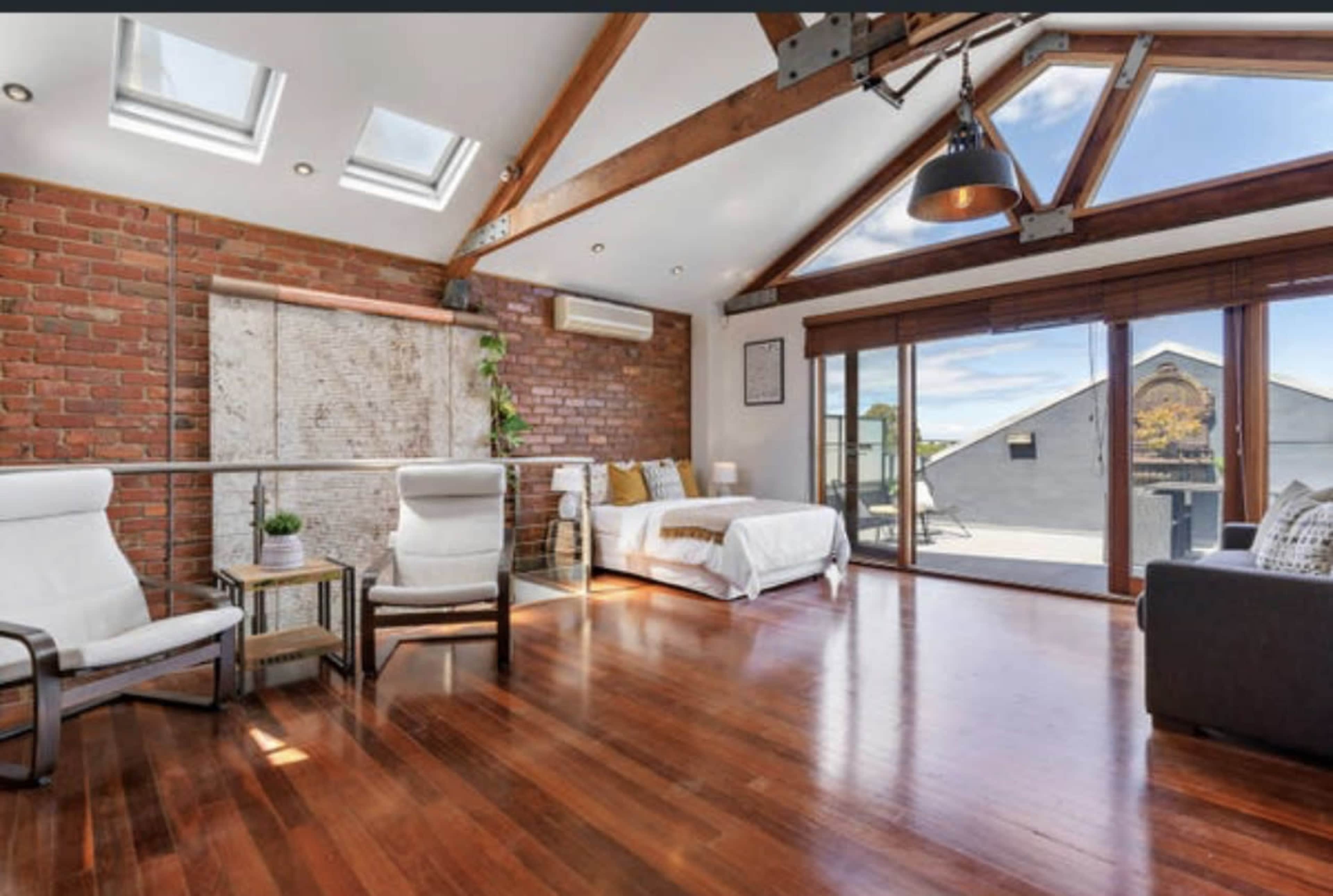A modern, airy room with a brick wall, wooden beams, large windows, and two white chairs facing a bed and a sliding glass door leading to a terrace.