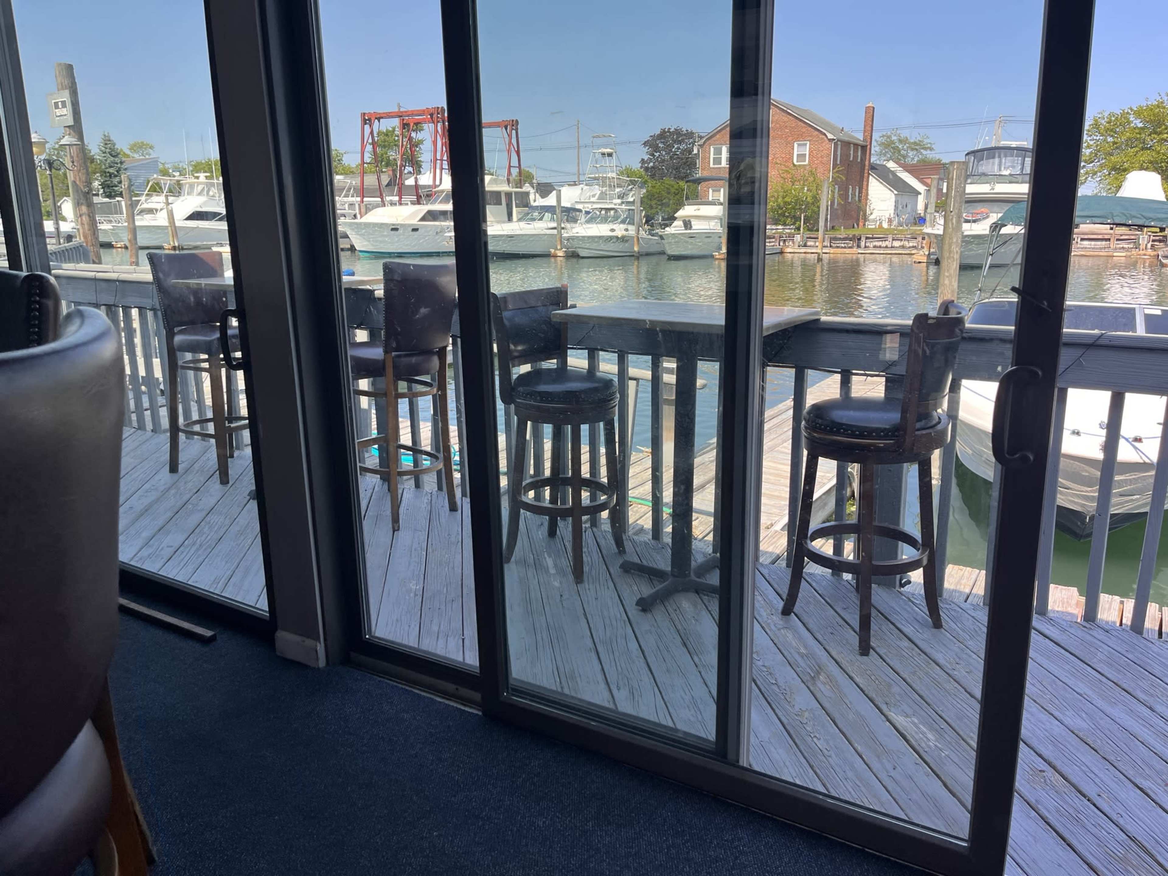 The image shows a view of a marina through large glass doors, with several bar stools positioned along a wooden deck overlooking the water and boats.