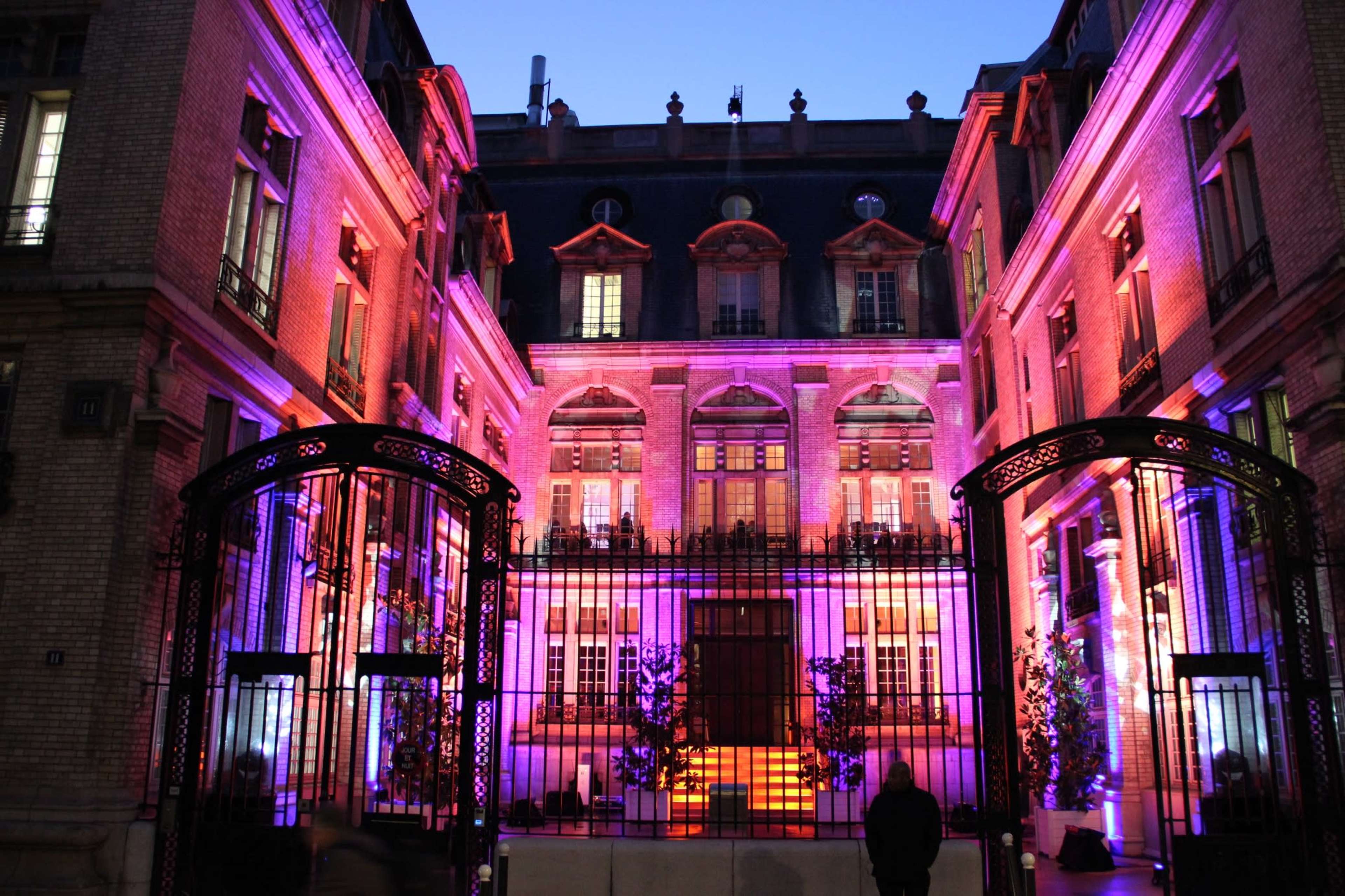 A historic building with a wrought iron gate is illuminated in purple and pink lights against a twilight sky.