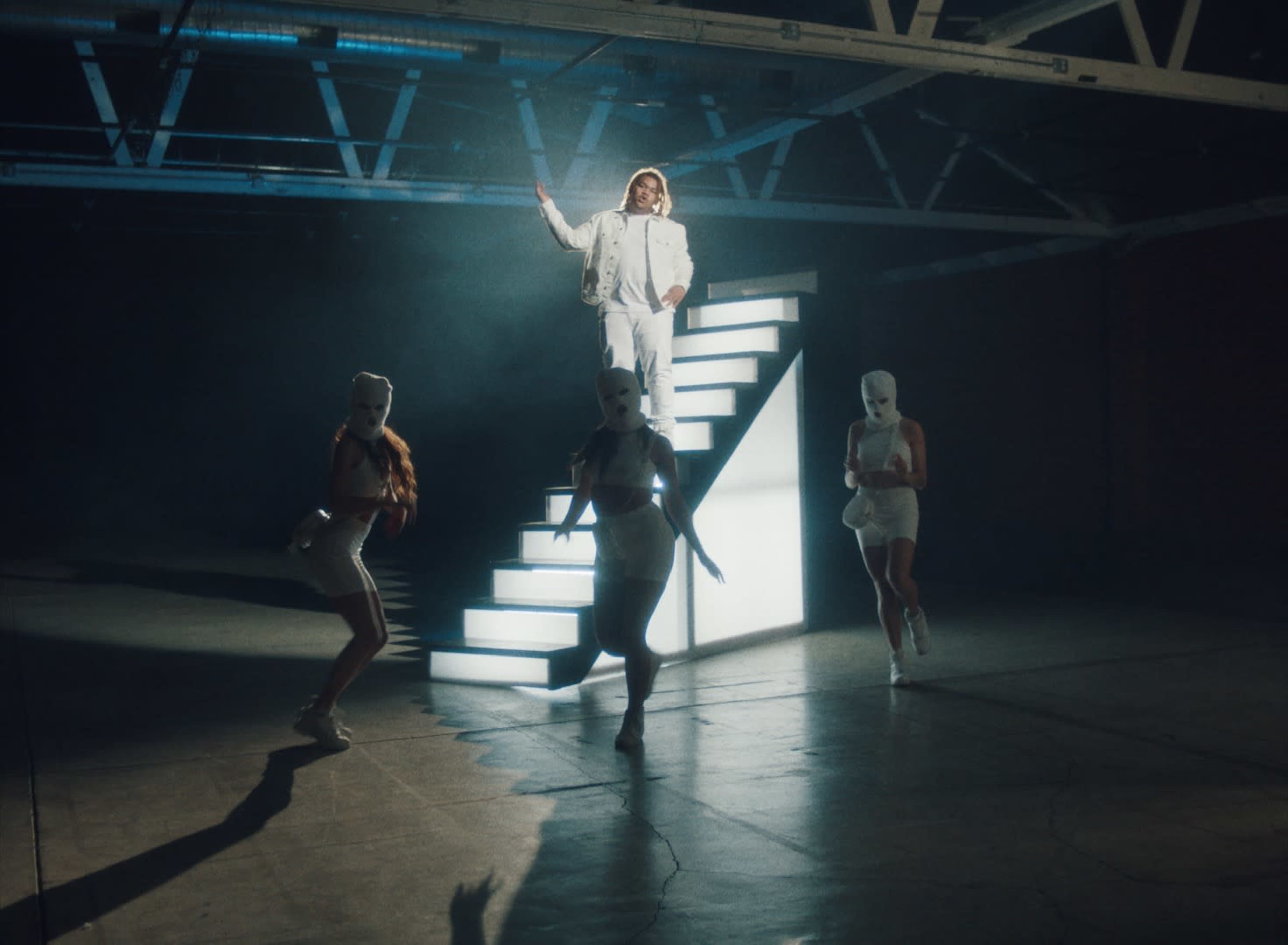 A performer dressed in white stands on illuminated stairs while three dancers in masks pose around them in a dimly lit warehouse.