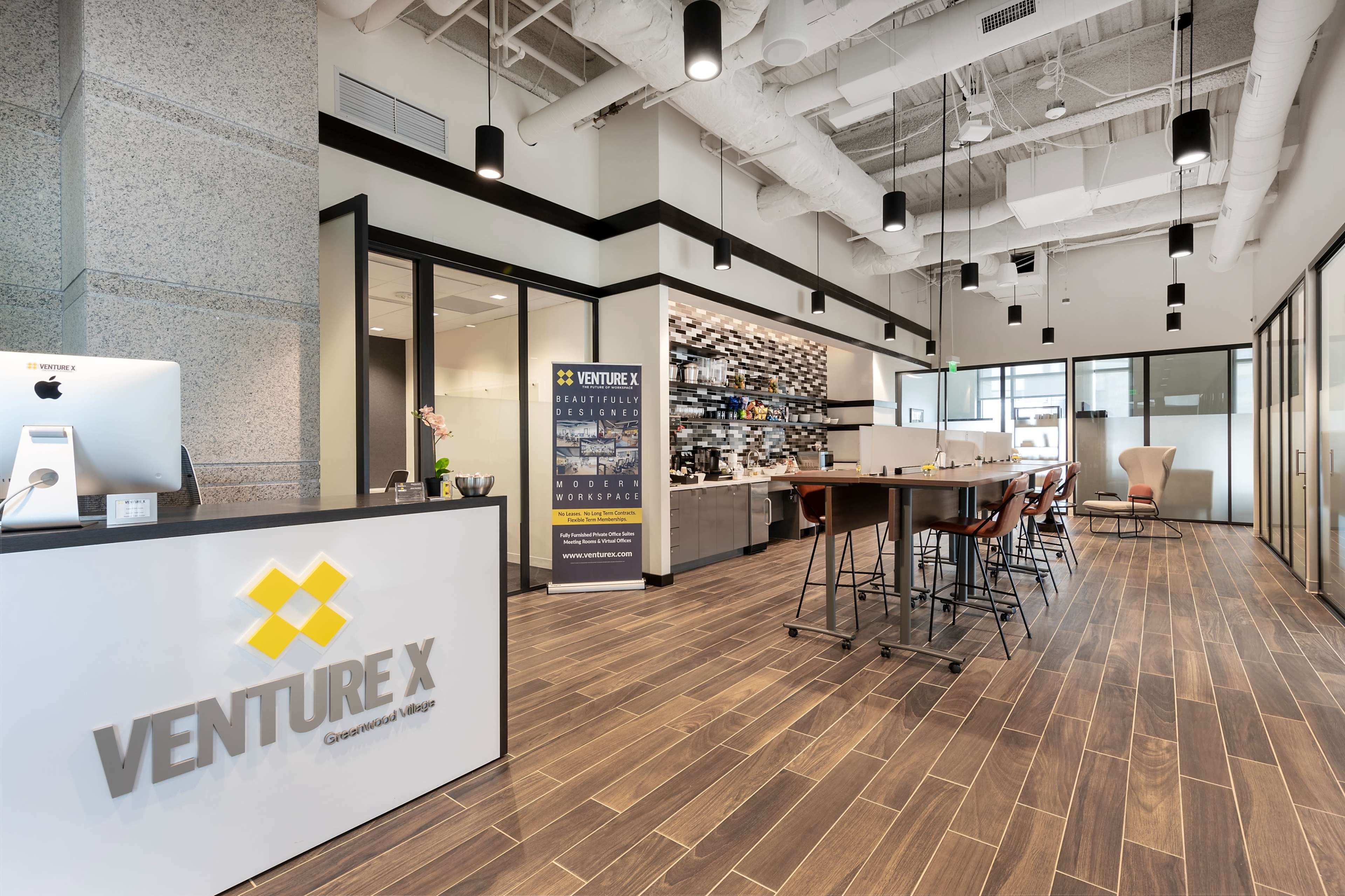 A modern coworking space featuring a reception area with a desk, a coffee station, high tables with stools, and glass-walled meeting rooms.