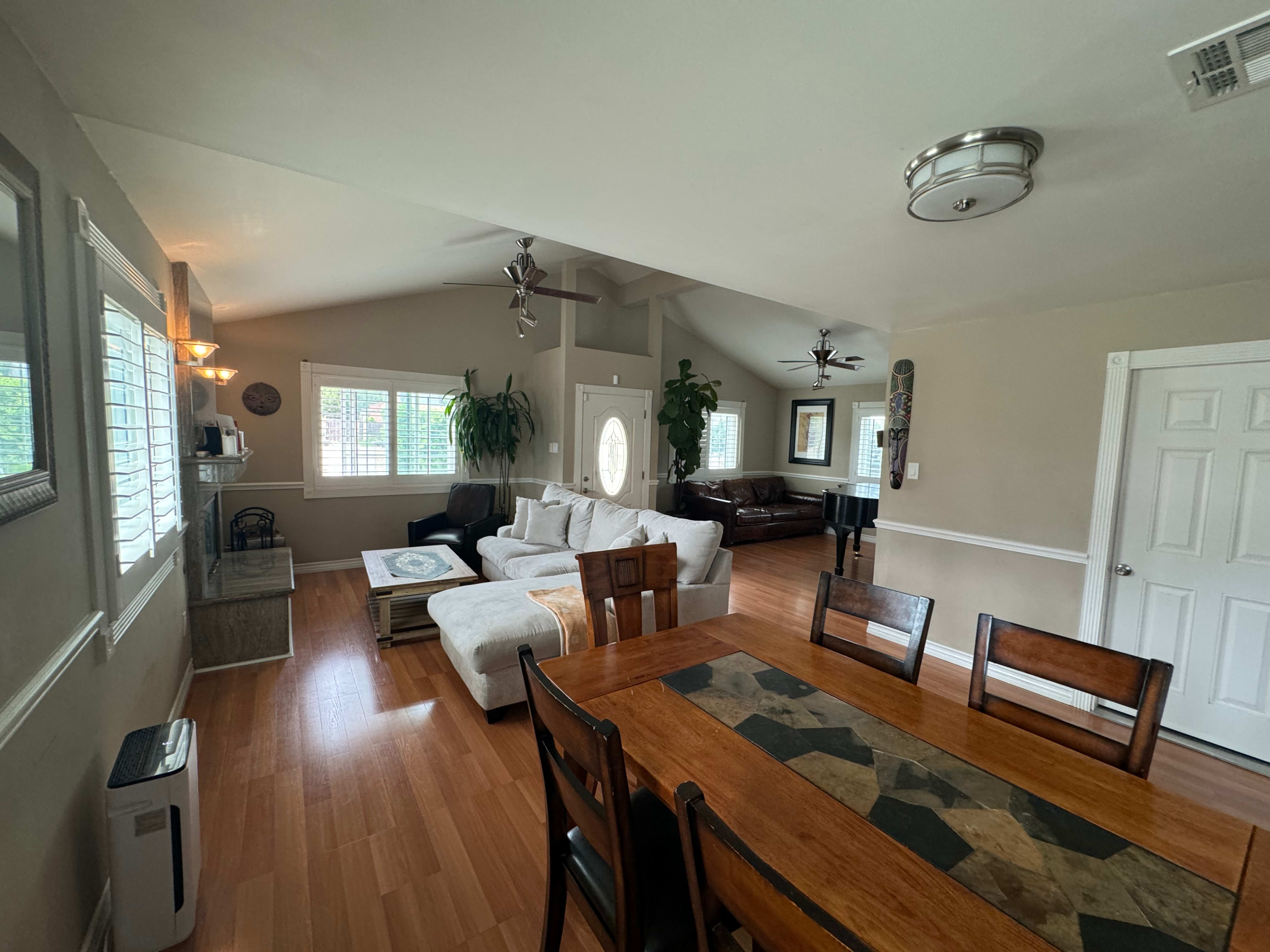 The image shows a spacious living area featuring a dining table, a cozy seating arrangement with a light-colored sofa, and a piano, all within a well-lit space.
