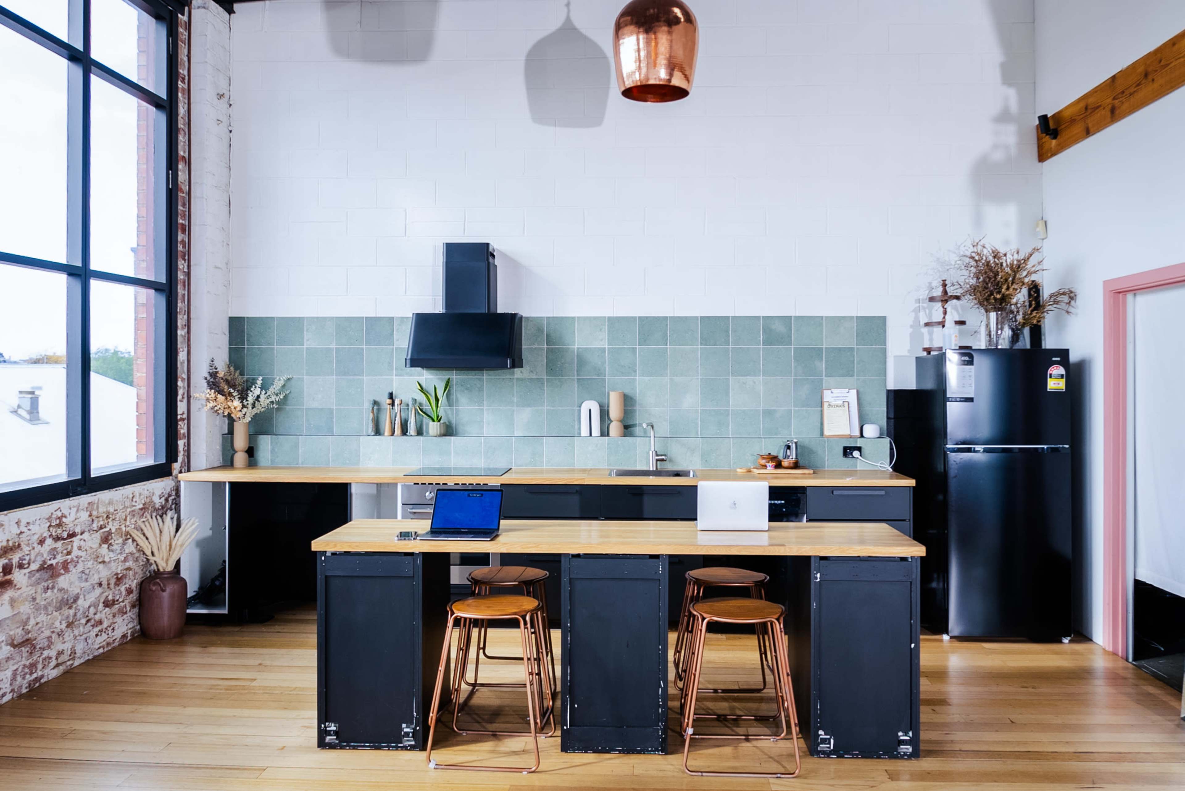A modern kitchen features a wooden island with two laptops on it, a green tiled backsplash, a black fridge, and large windows allowing natural light to enter.