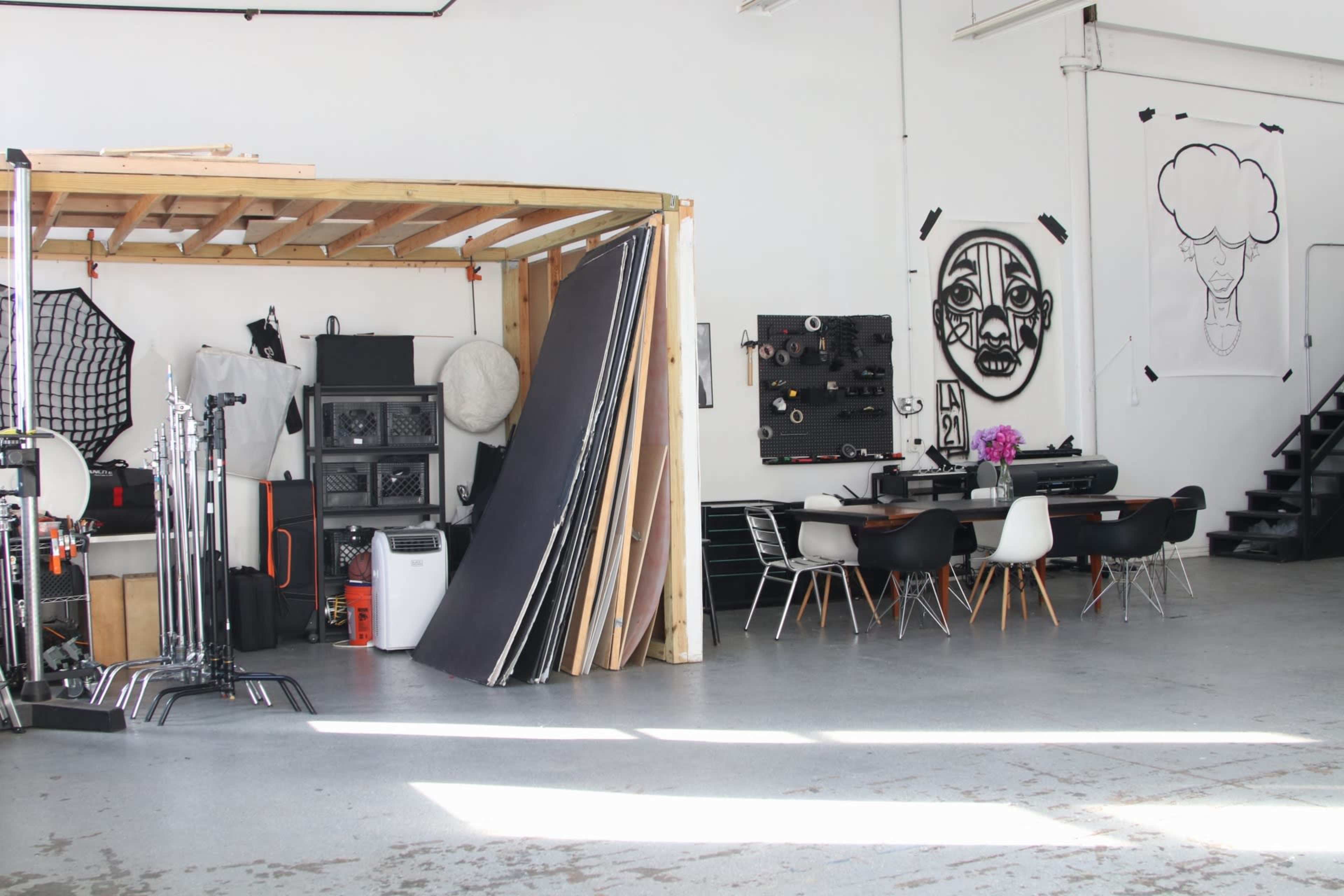 The image shows a spacious studio setup with wooden structures, various photography equipment, a large table surrounded by chairs, and wall art.