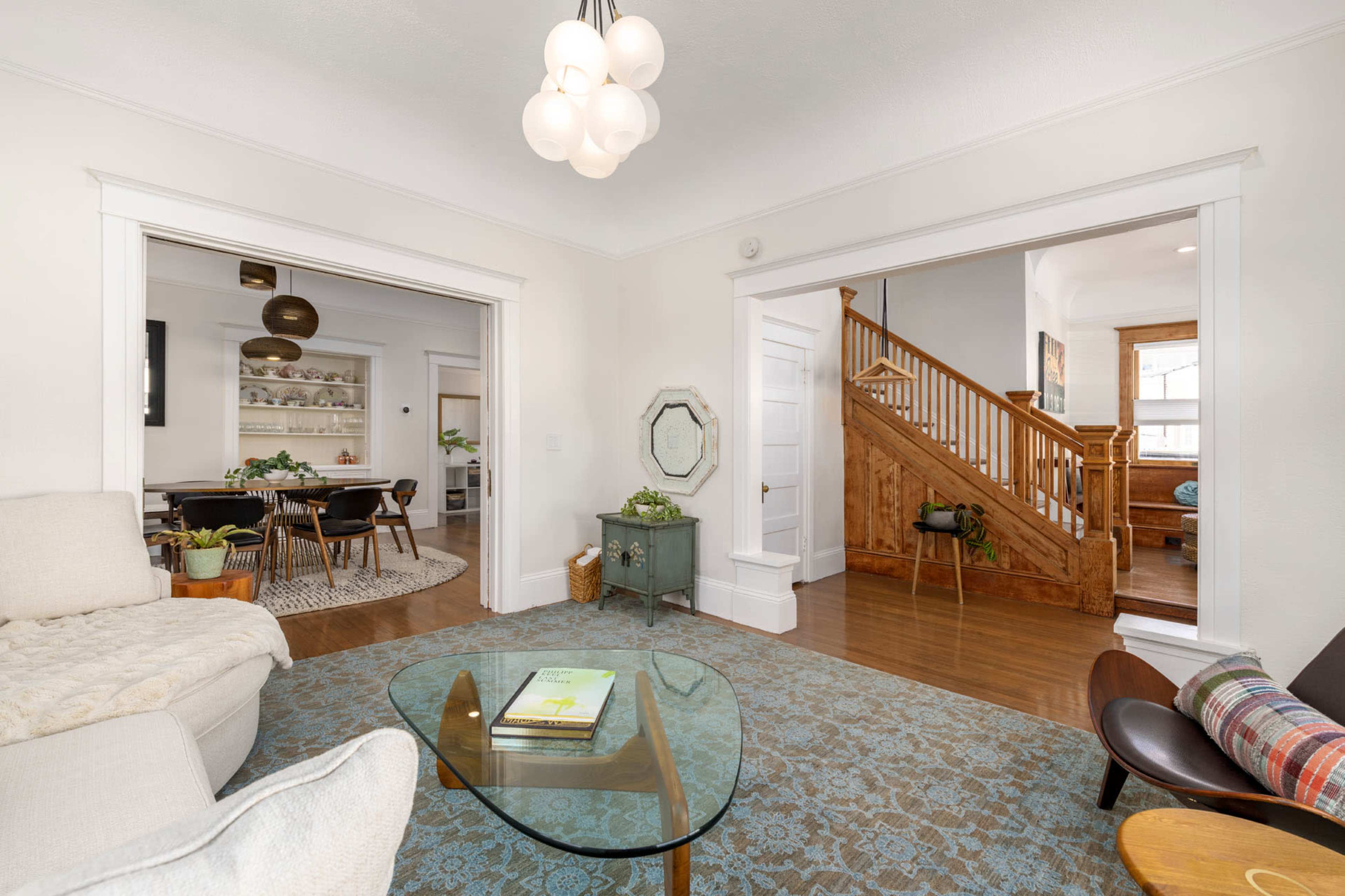 A well-lit living room with a light-colored sofa, a glass coffee table on a patterned rug, and views of an adjacent dining area and staircase.