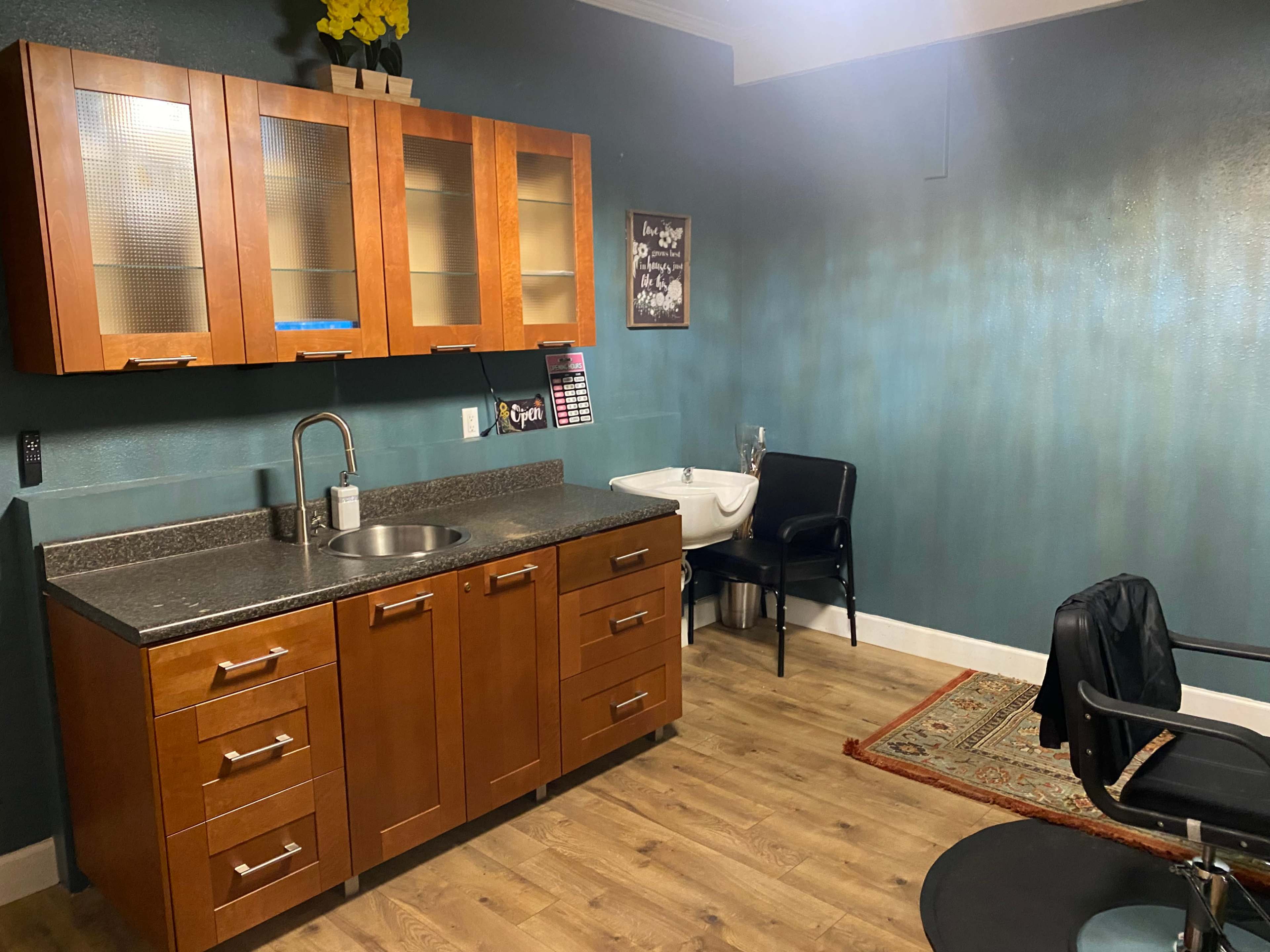 A small room features a wooden cabinetry with a sink, a black chair, and a decorative rug on a wood floor.