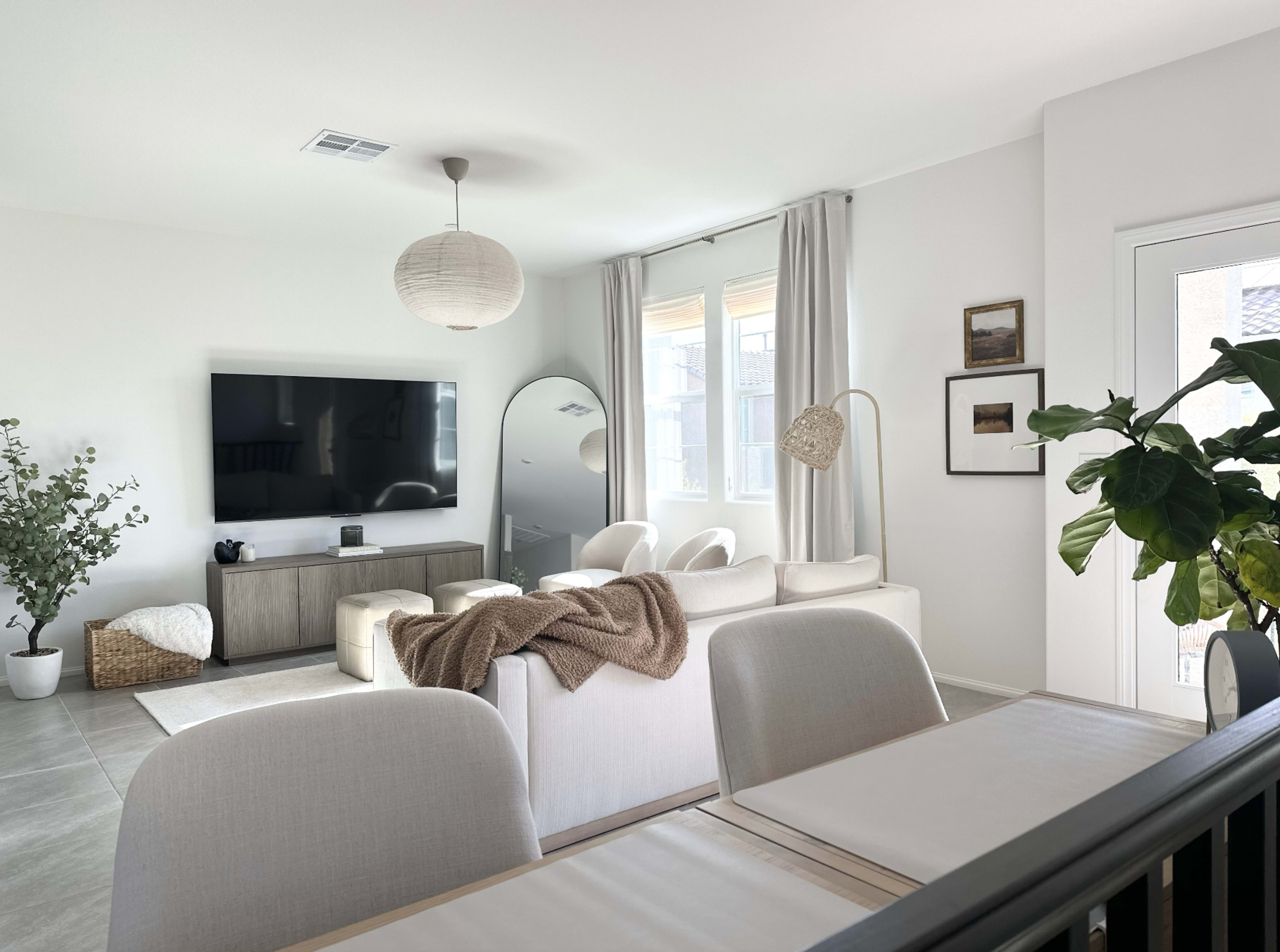 A modern living room features a large TV mounted on the wall, a sectional sofa, a round mirror, and bright windows, with decorative plants and light-colored furnishings.